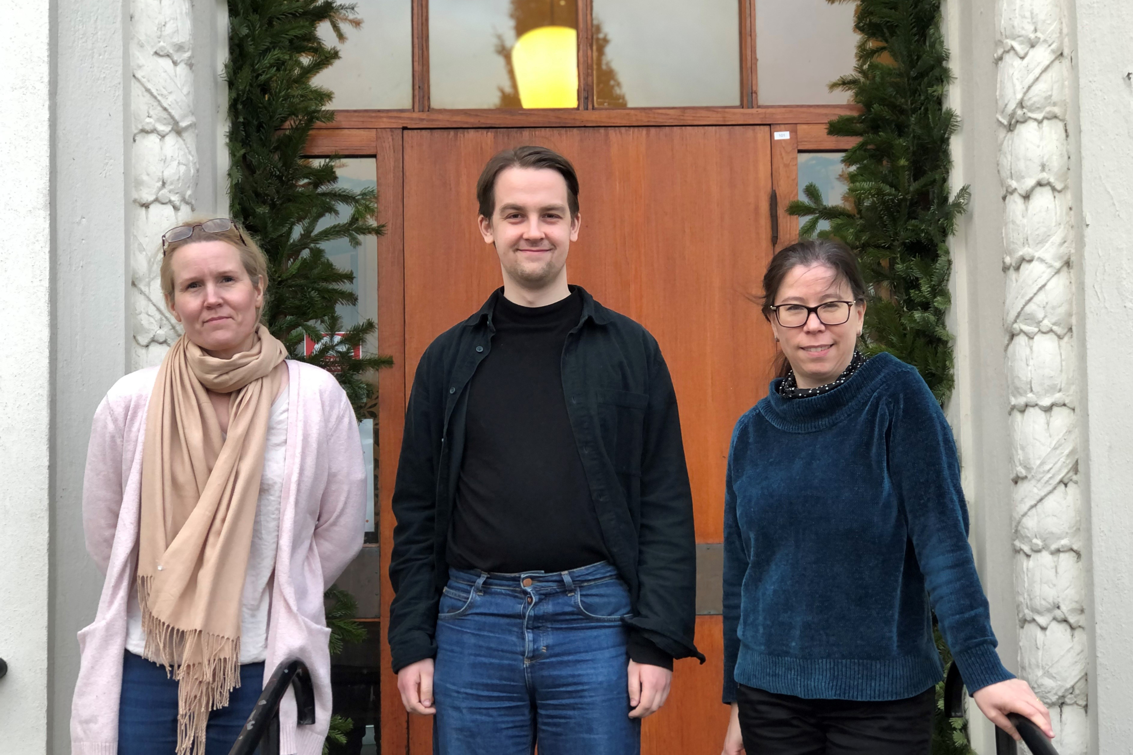 Blond person i ljusrosa kofta, mellanblond person i svart tröja och mörkhårig person i blå tröja på trappan till församlingshuset