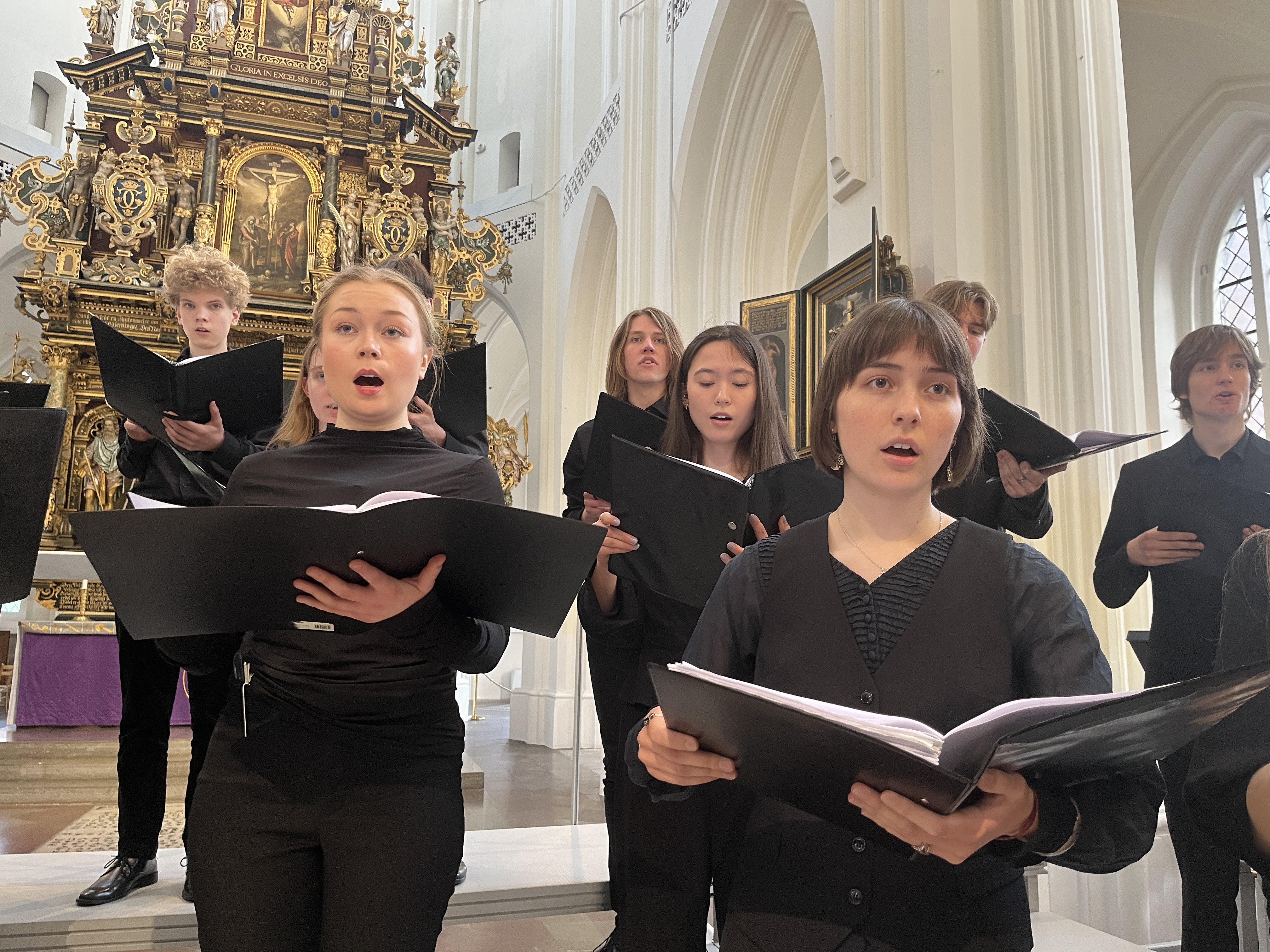 Grupp av unga kvinnor och män klädda i svart håller i svarta pärmar och sjunger i en kyrka.