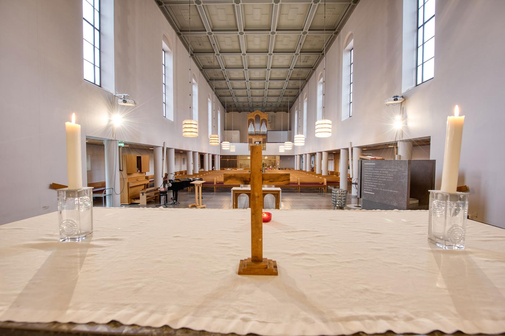 Närbild på altare med vit duk, ljus och kors. Framför alltaret breder ett avlångt kyrkorum ut sig med vita väggar, höga fönster och bänkar i trä.