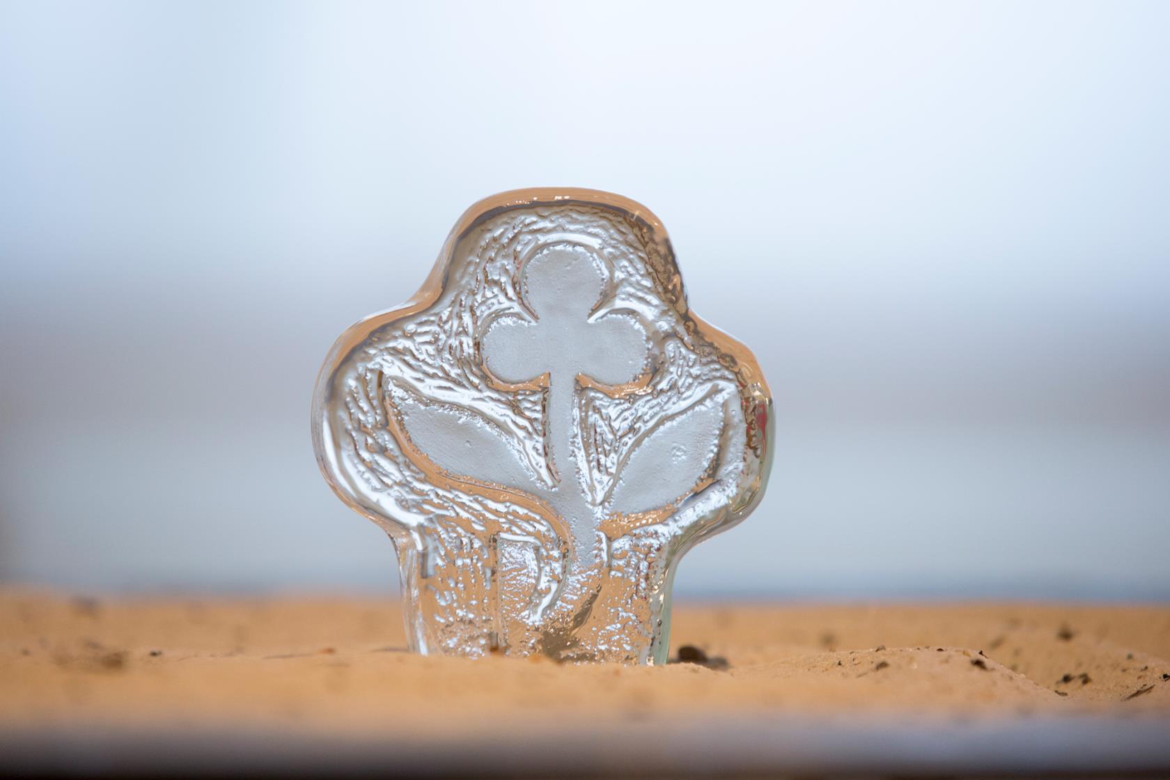 En glasskulptur föreställande en blomma innesluten i ett kors står på en bädd av sand.