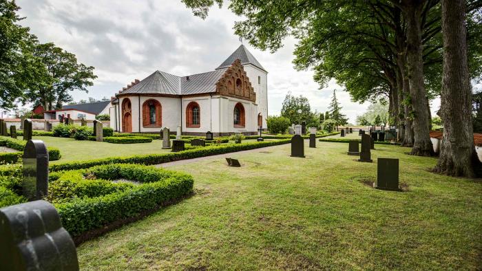 Vitputsad kyrka med röda tegeldetaljer omgiven av en lummig kyrkogård.