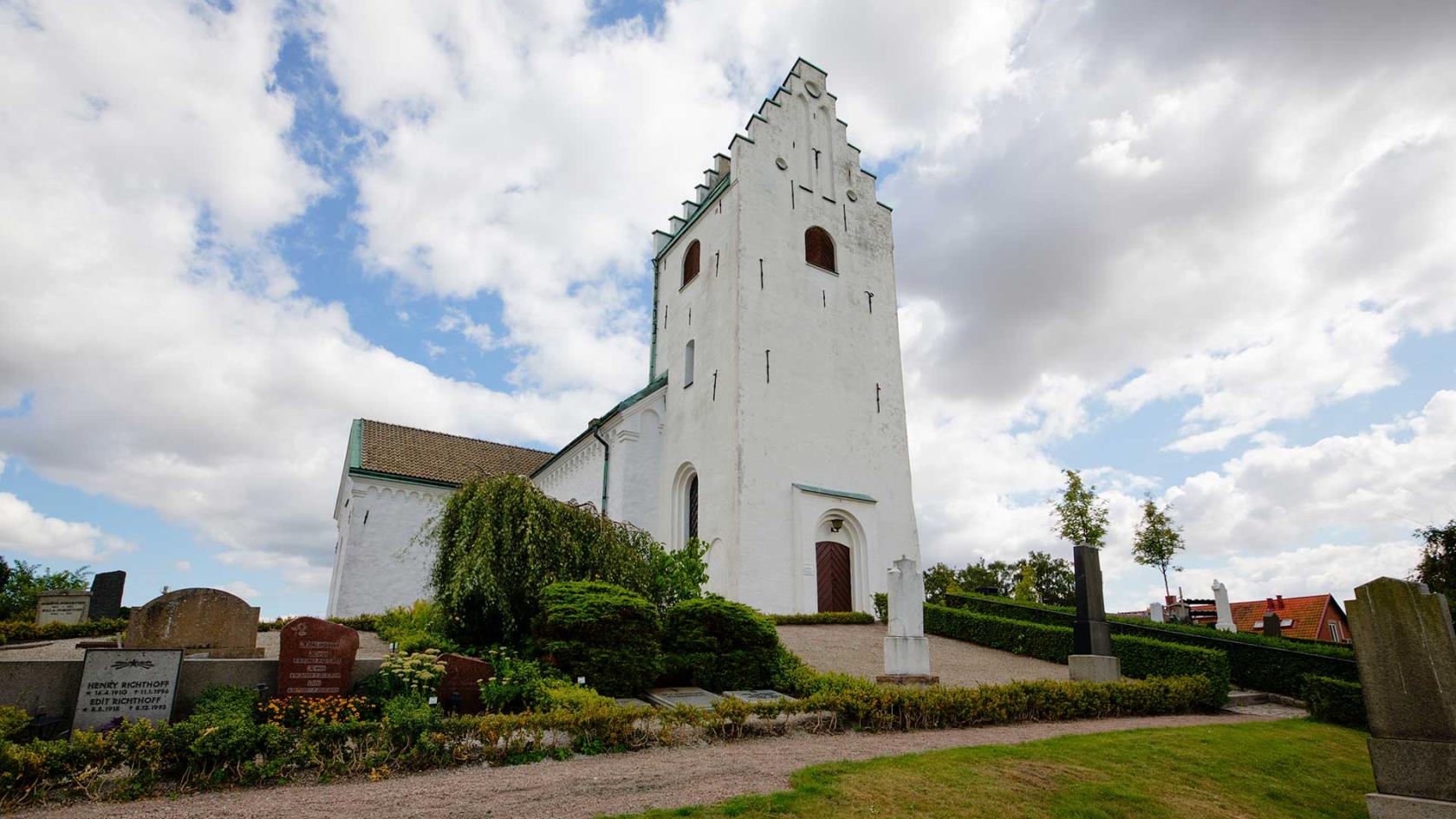 Vitkalkad kyrka med torn med trappstensgavel ligger uppe på en kulle. Runt om finns gravstenar, gräs och grusgångar.