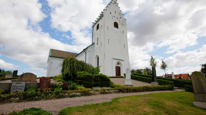 Vitkalkad kyrka med torn med trappstensgavel ligger uppe på en kulle. Runt om finns gravstenar, gräs och grusgångar.