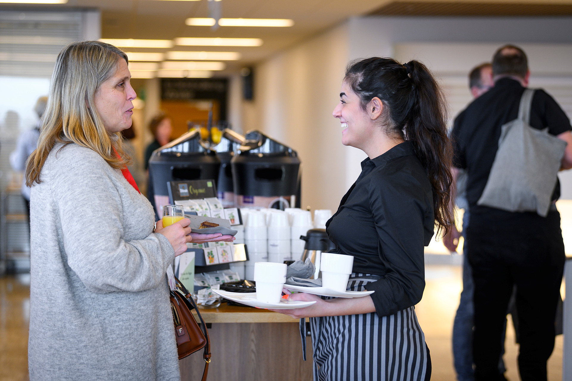 Två kvinnor står och pratar och skrattar, den yngre kvinnan bär på en bricka med koppar och fat.. Den ena är lite äldre än den andra. I bakgrunden syns en man bakifrån.