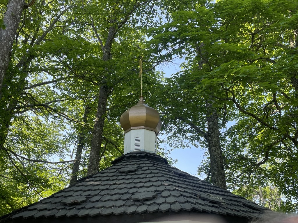 Tornspira i guld/vitt på ett litet kapell med svart tak. Grönt runt omkring av trädkronor.