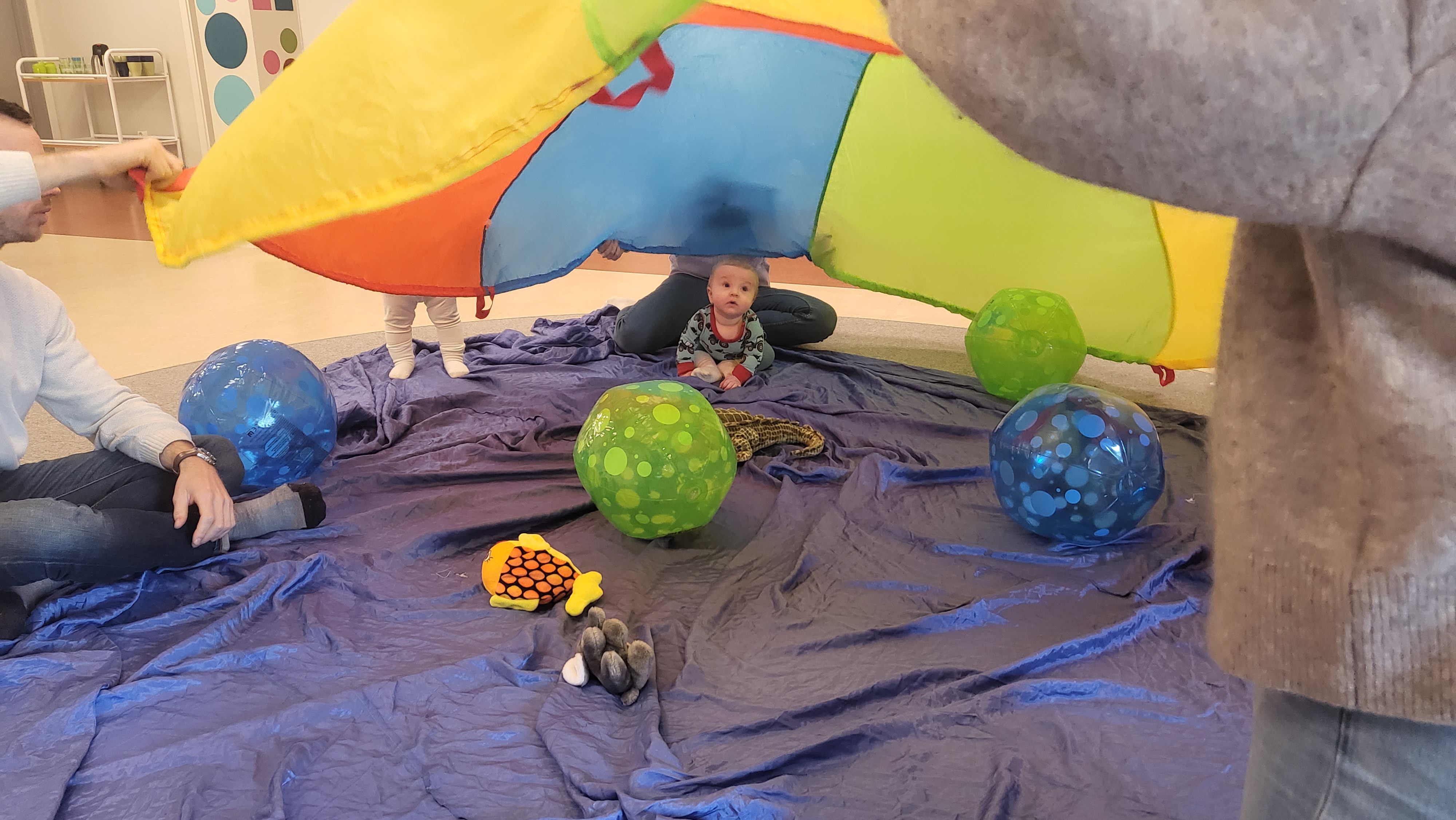 Ett blått stort skynke som symboliserar havet ligger på golvet. Tillsammans sitter barn och föräldrar i en ring. De vuxna håller i en färgglad fallskärm som flyger upp och ner över barnen som ligger eller sitter på det blå skynket. På skynket finns även gosedjur och färgglada badbollar i grönt och blått.