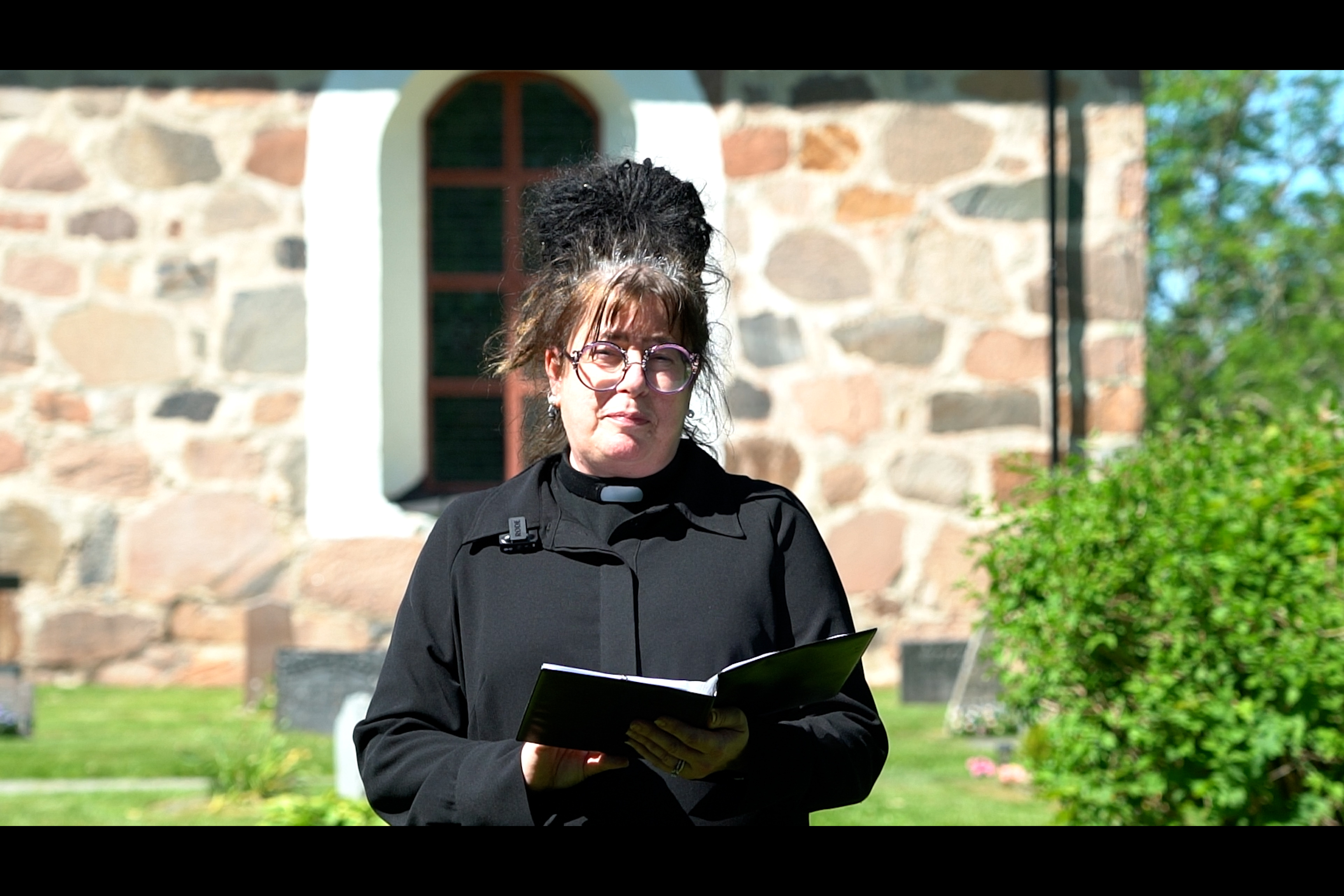 En kvinna i svart, iklädd prästkrage, står framför en kyrka i solen.