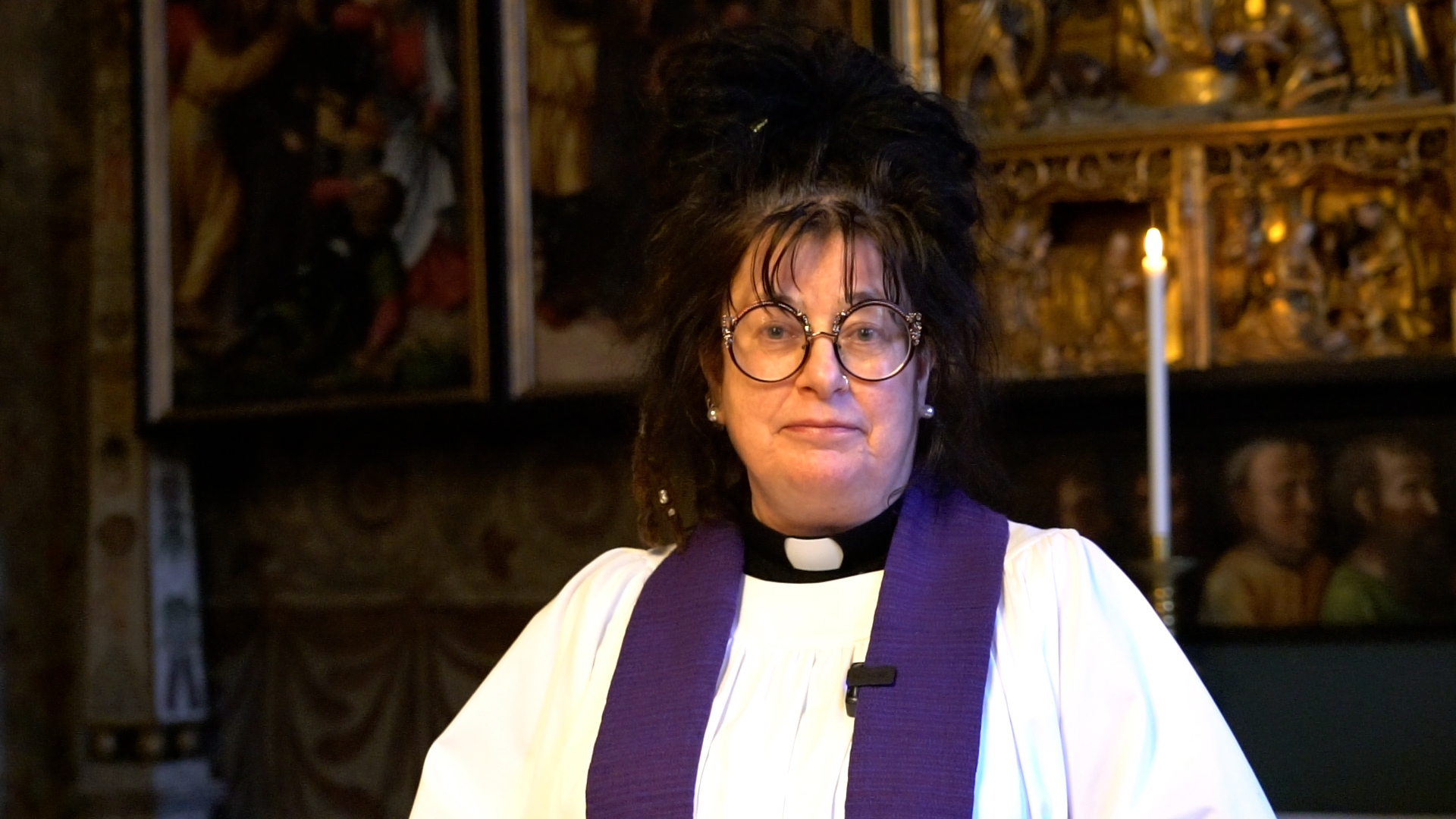 En kvinnlig präst med stort svart hår, stora guldglänsande glasögon och vit klädnad med lila stola, står framför ett guldfärgat altare. 