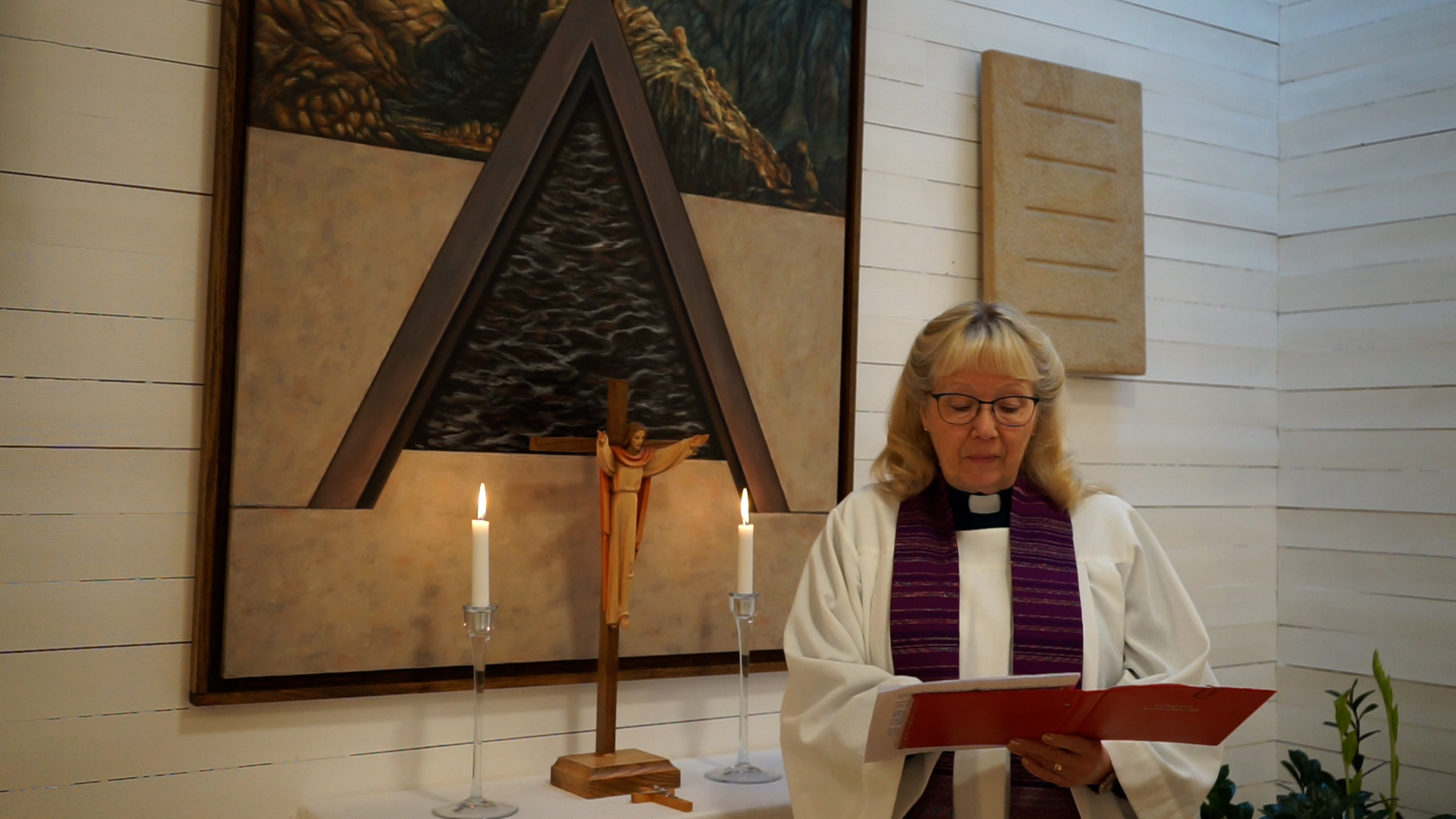 En kvinnlig präst står framför ett altare där två ljus brinner. Prästen är klädd i vit klädnad och en lila stola. 