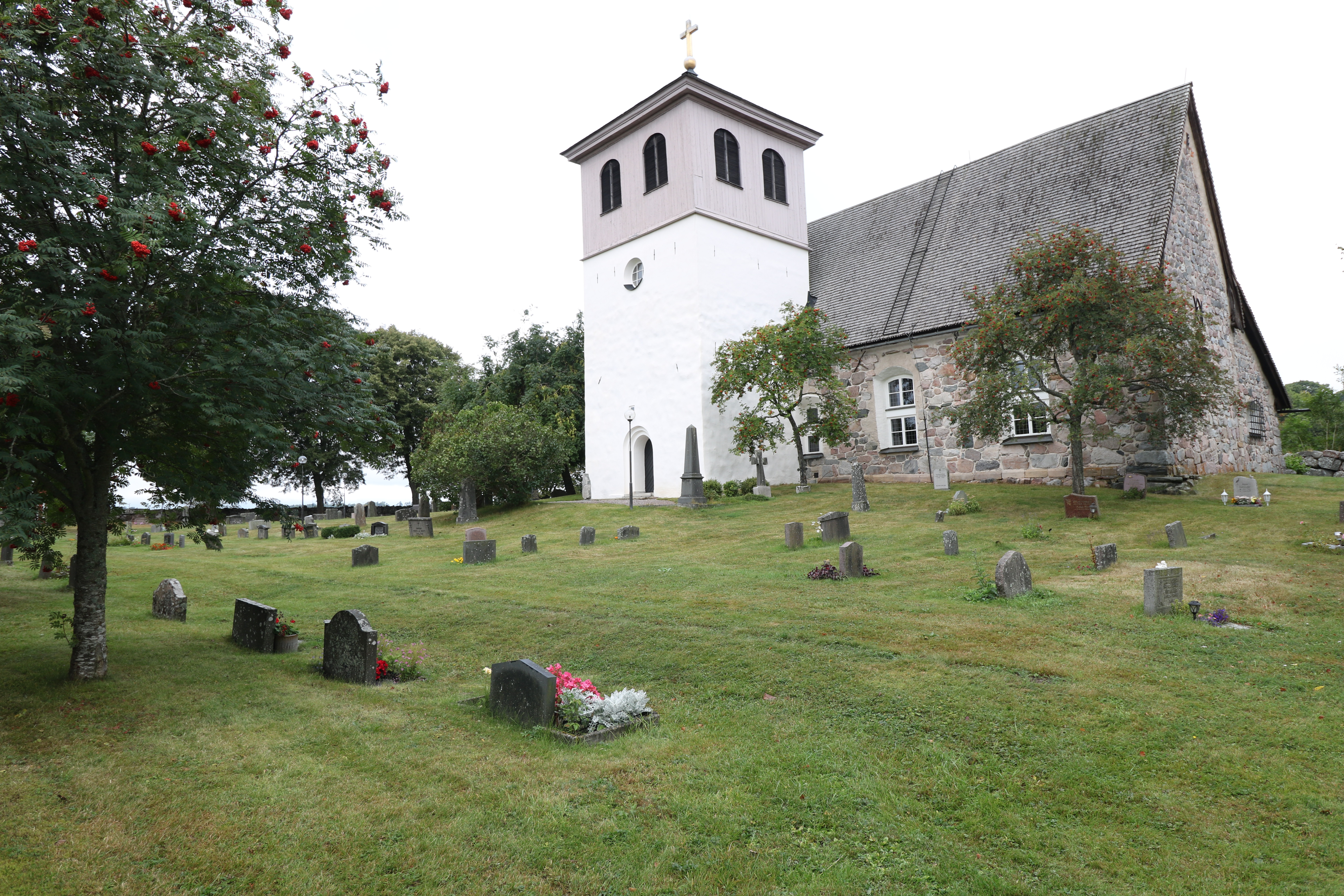 En stenkyrka med vitt kyrktorn i bakgrunden. Framför kyrkan kyrkogården med grönt gräs och utspridda gravstenar.