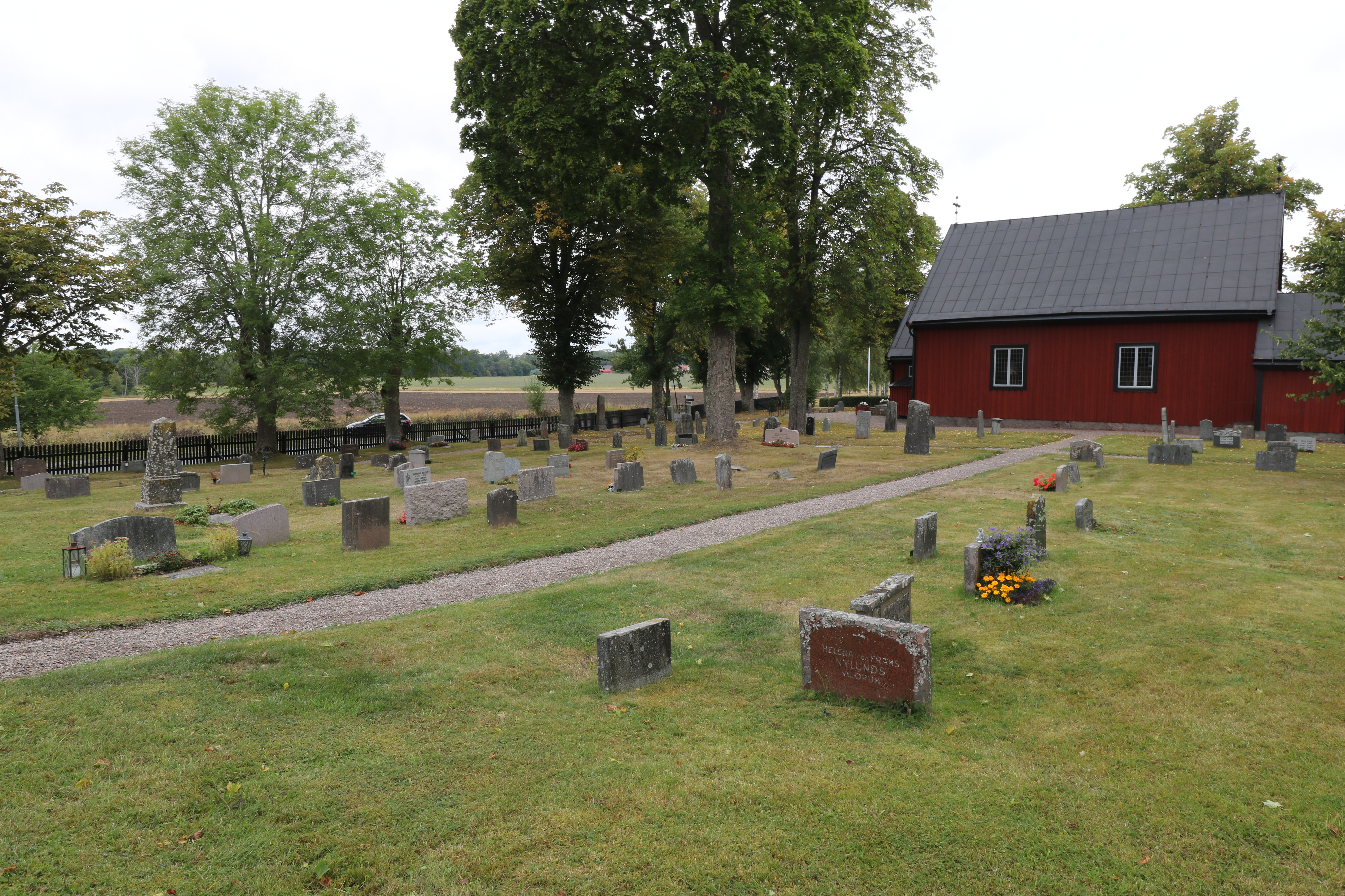 Gravstenar på en kyrkogård. Ett kapell med röda väggar och ett svart tak längre bak i bilden.