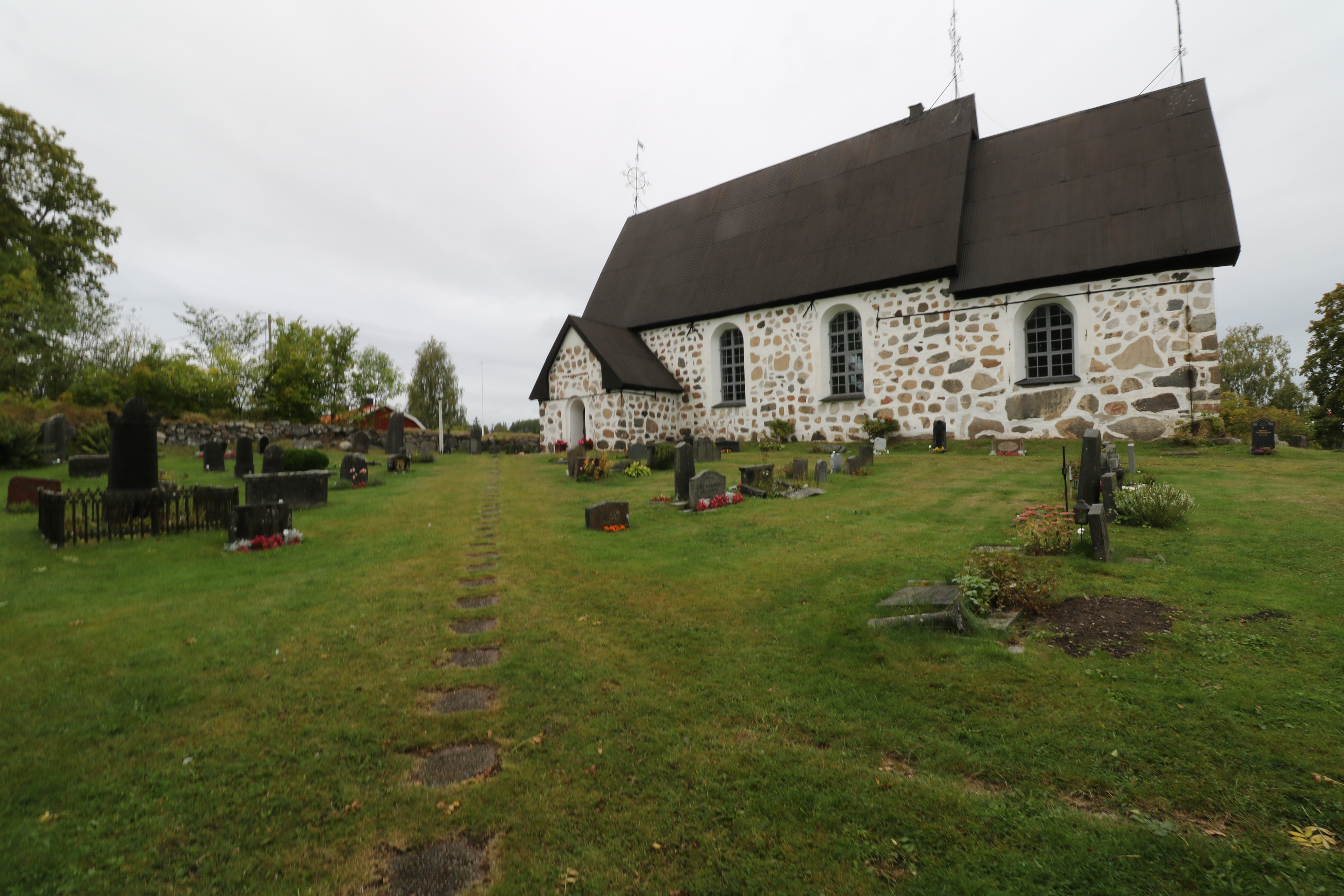 En sten kyrka i vitt med gravstenar framför i gräsmattan