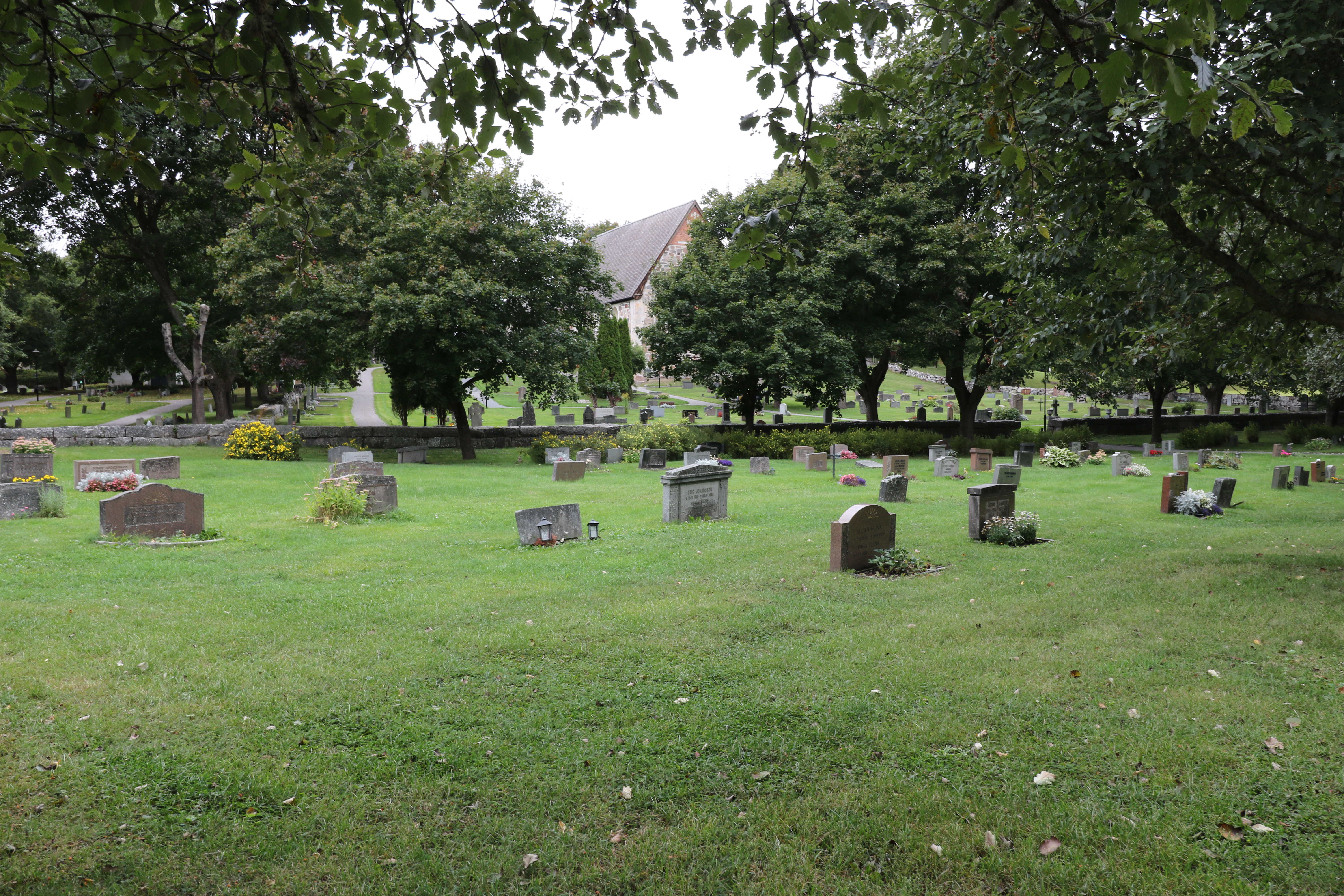 En stor gräsmatta med gravar och träd, långt bort skymtar en kyrka mellan träden.