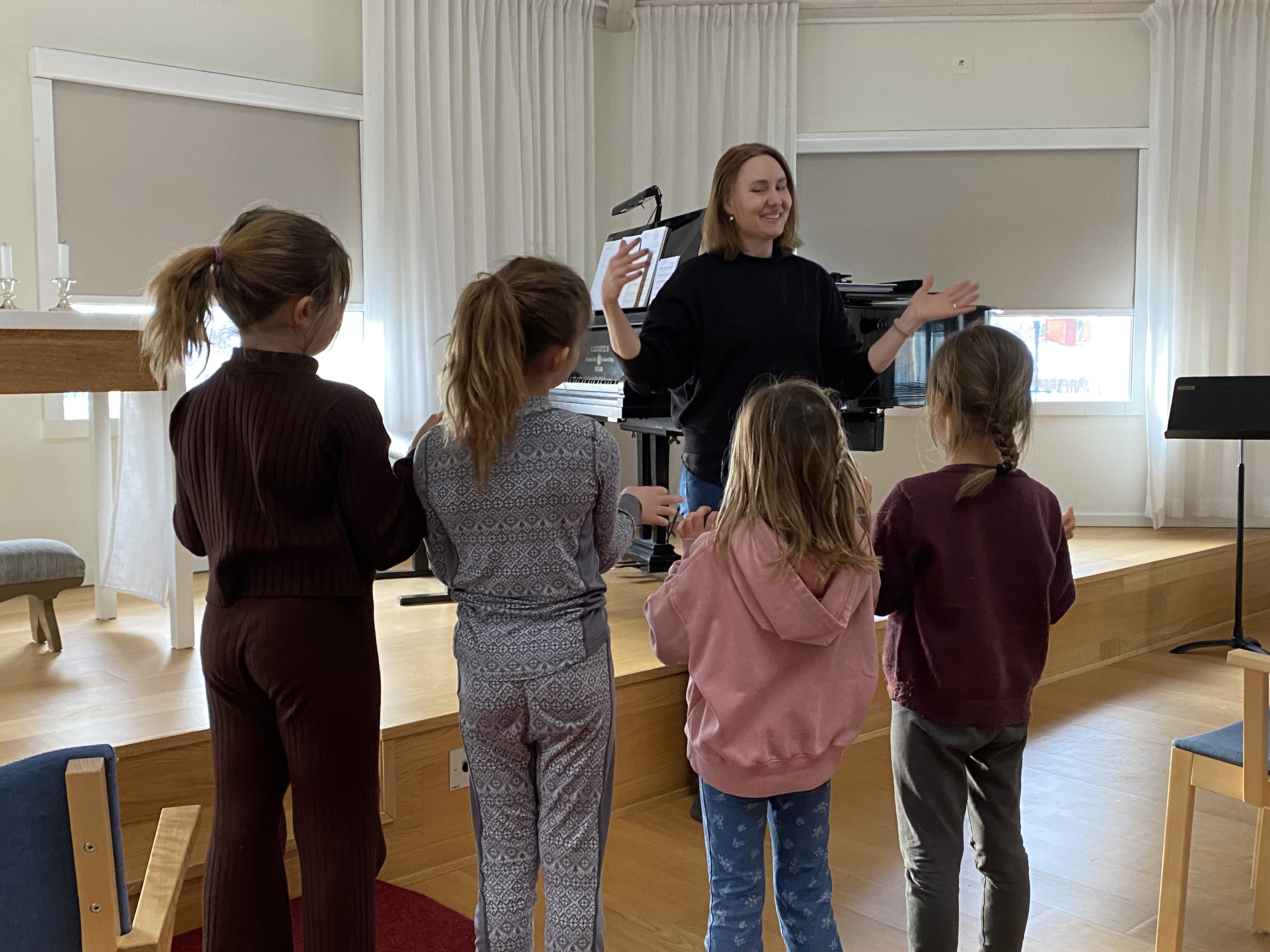 Några barn står med ryggarna mot kameran och tittar på en kvinna som viftar med handen. 