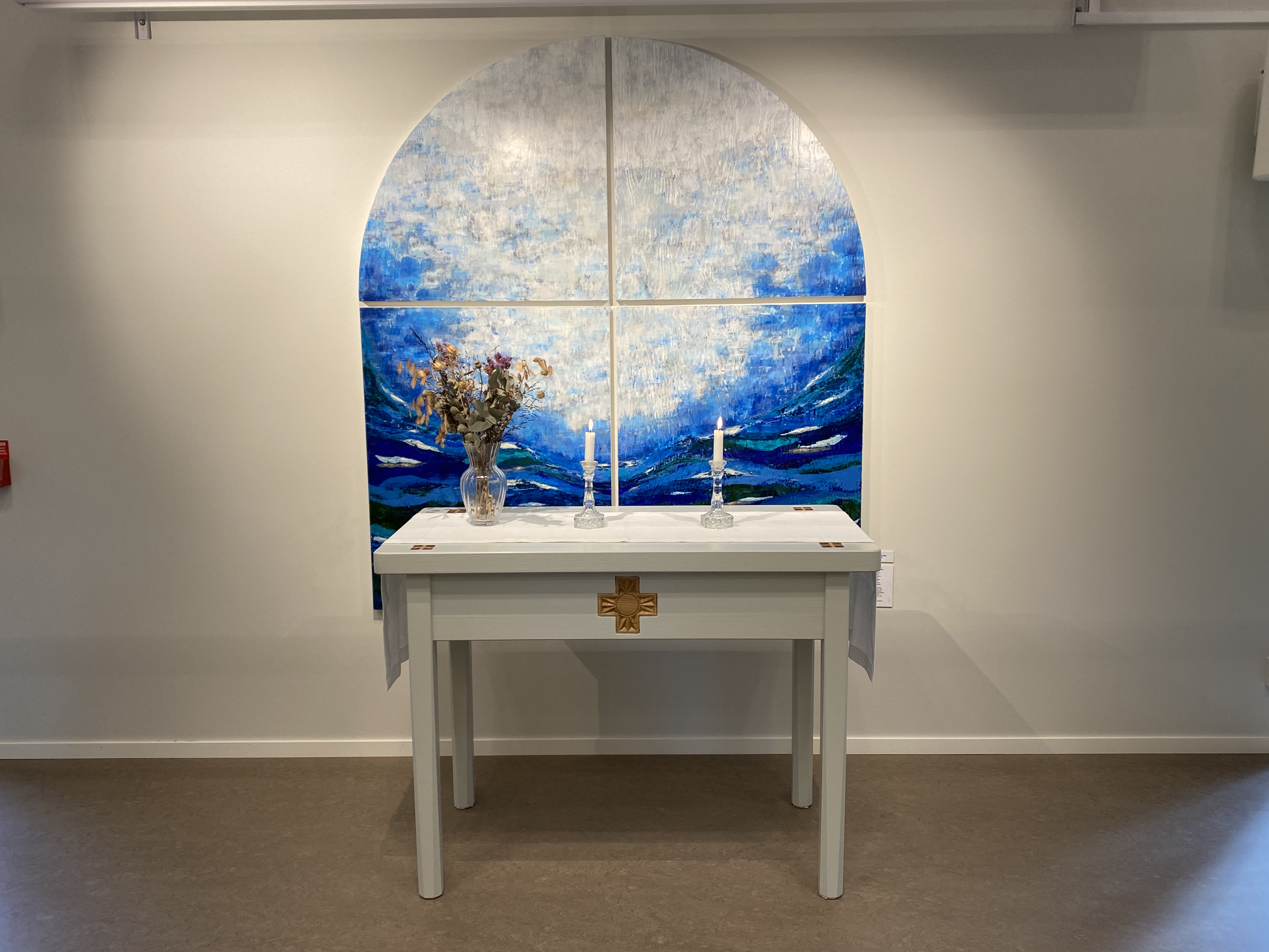Ett vitt altare står framför en fyrdelad målning med himmel och hav i form av ett kyrkfönster.