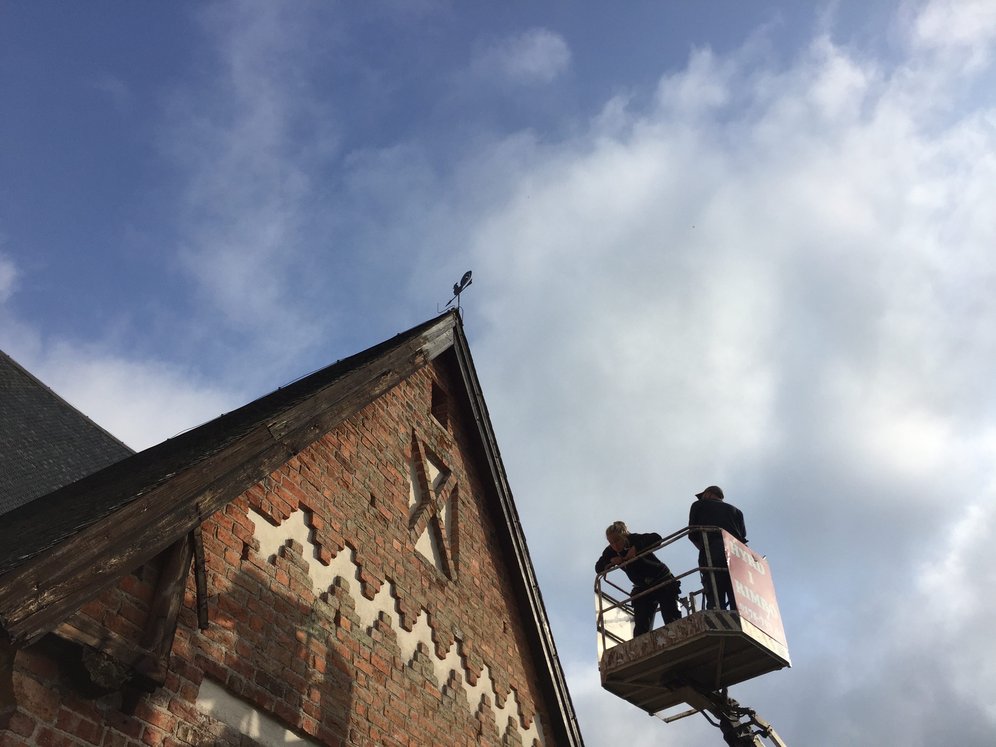 Två personer i en skylift på väg upp mot nocken på en kyrka. 