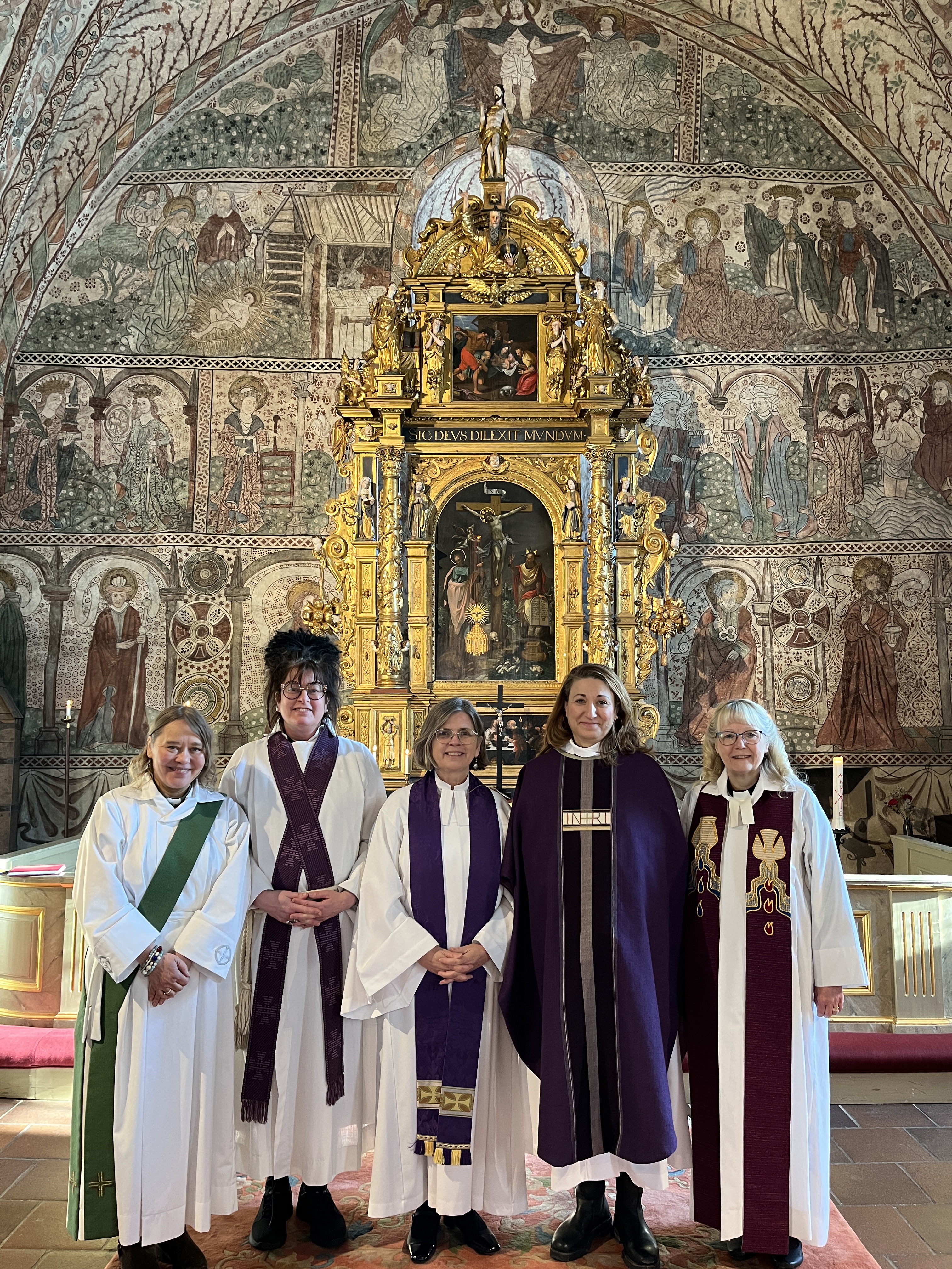 Fem personer står framför ett guldskimrande altare. Fyra är präster och en är en diakon.