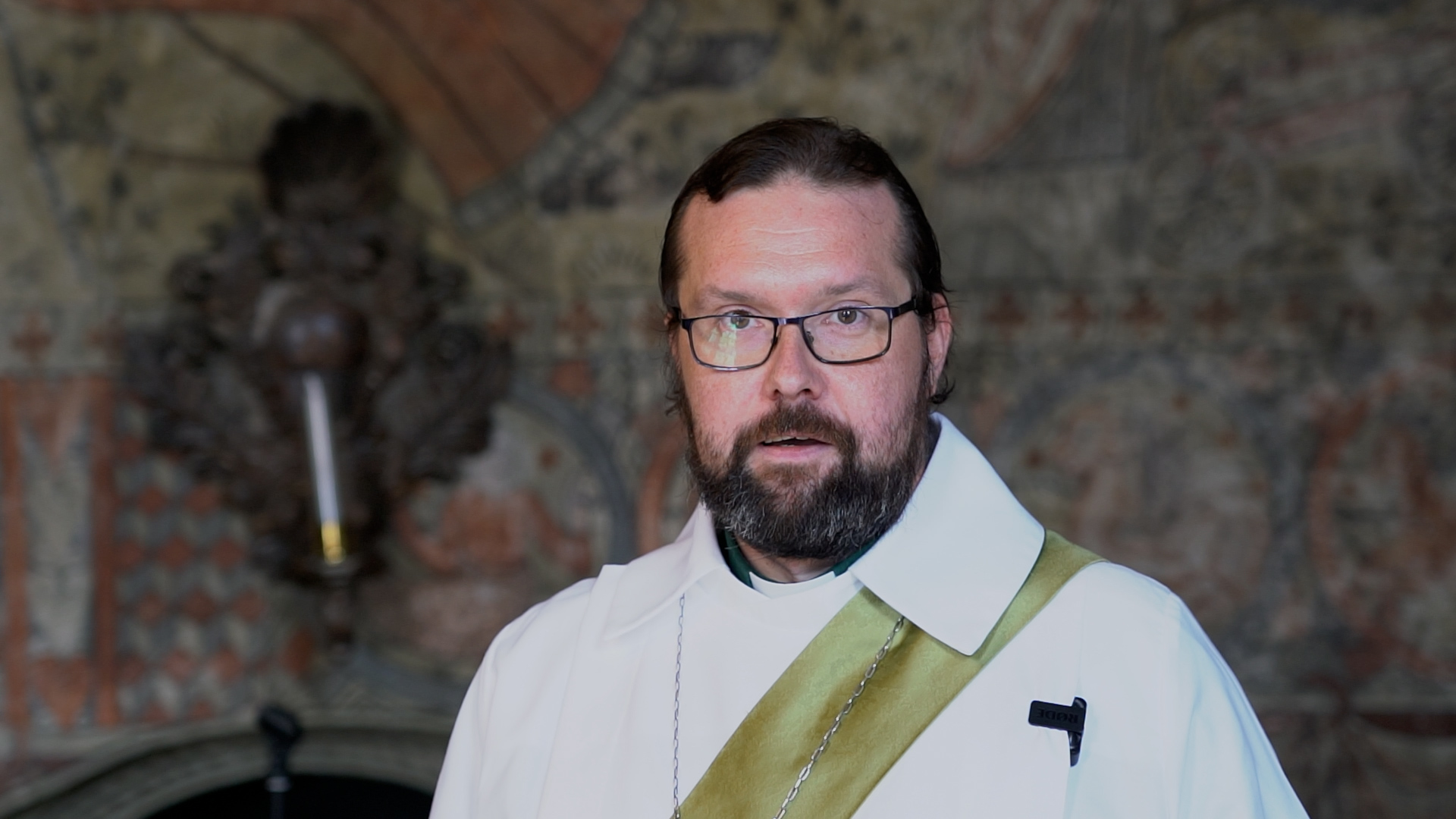 En man i en vit klädnad med en grön stola. Mannen har mörkt skägg och glasögon.