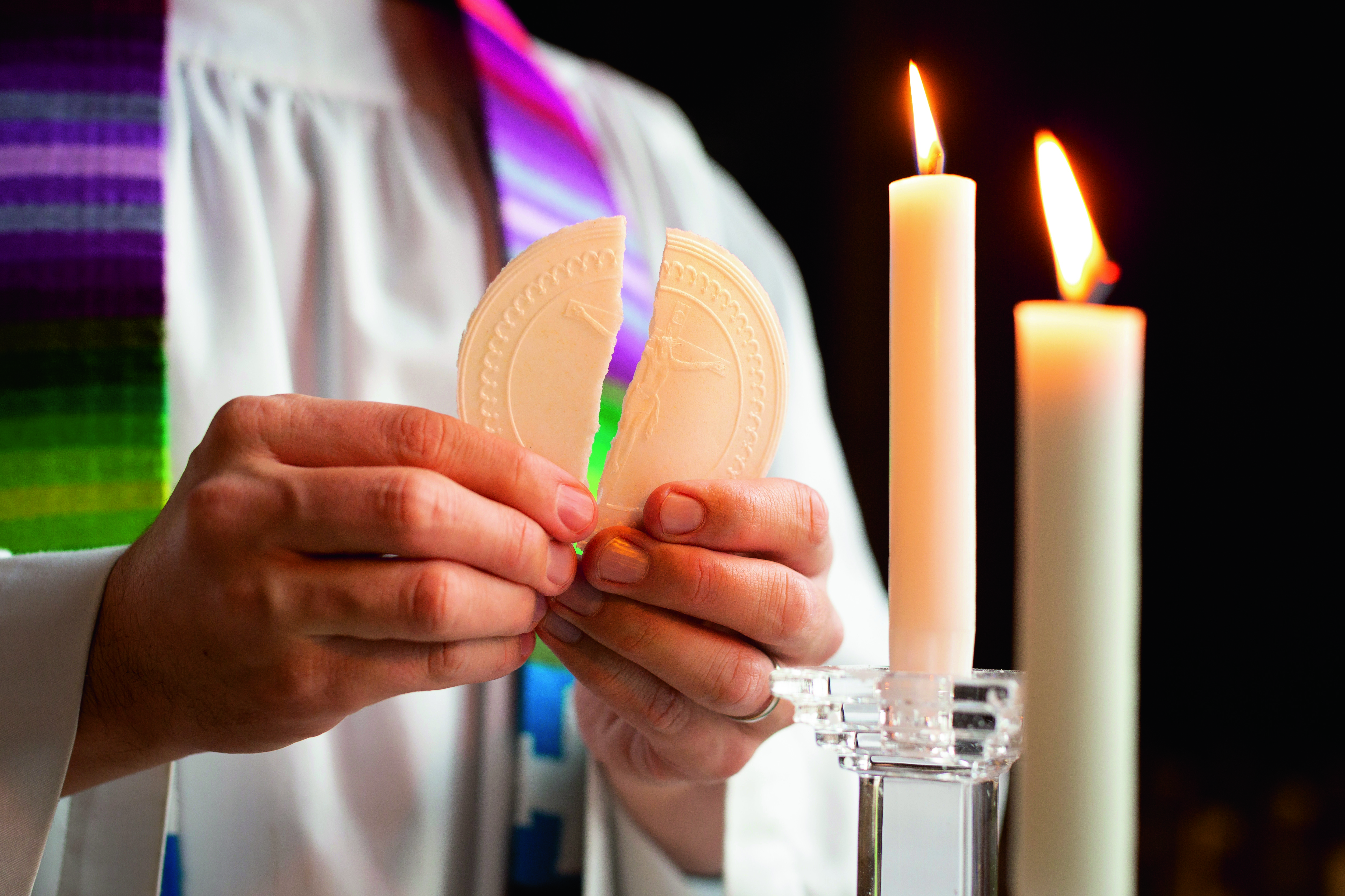 Två händer som bryter en oblat, i bakgrunden två brinnande ljus.