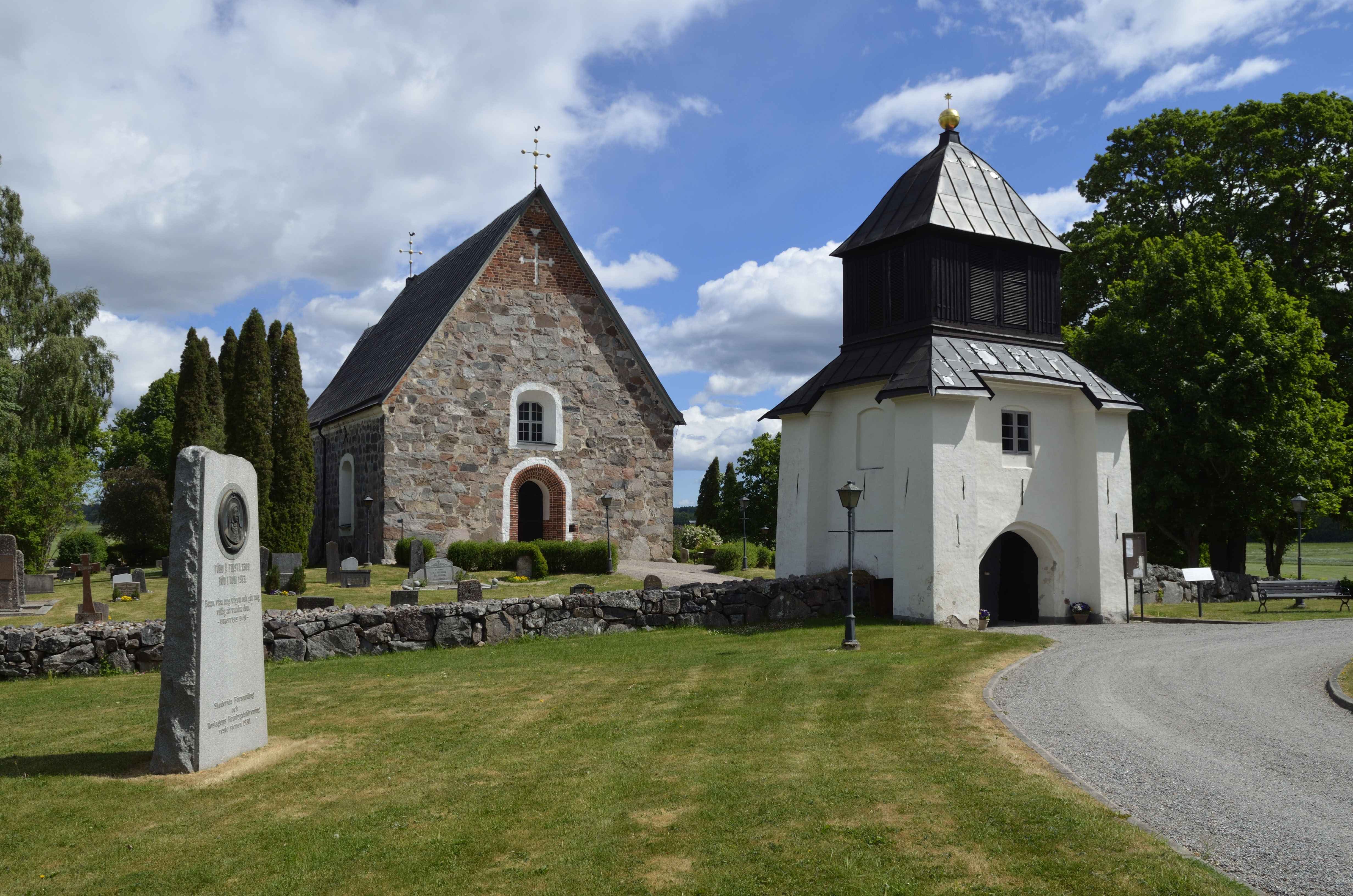 En minnessten står framför en brun stenkyrka. Man ser också en vit stiglucka som är ingången till kyrkogården.