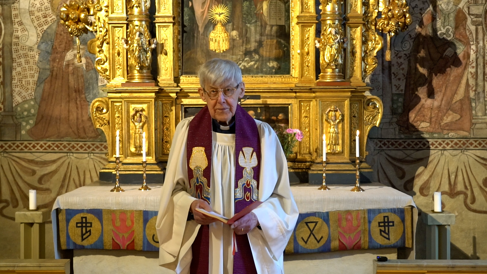 En präst i vit klädnad med en stola i lila står framför ett guldskimrande altare. 