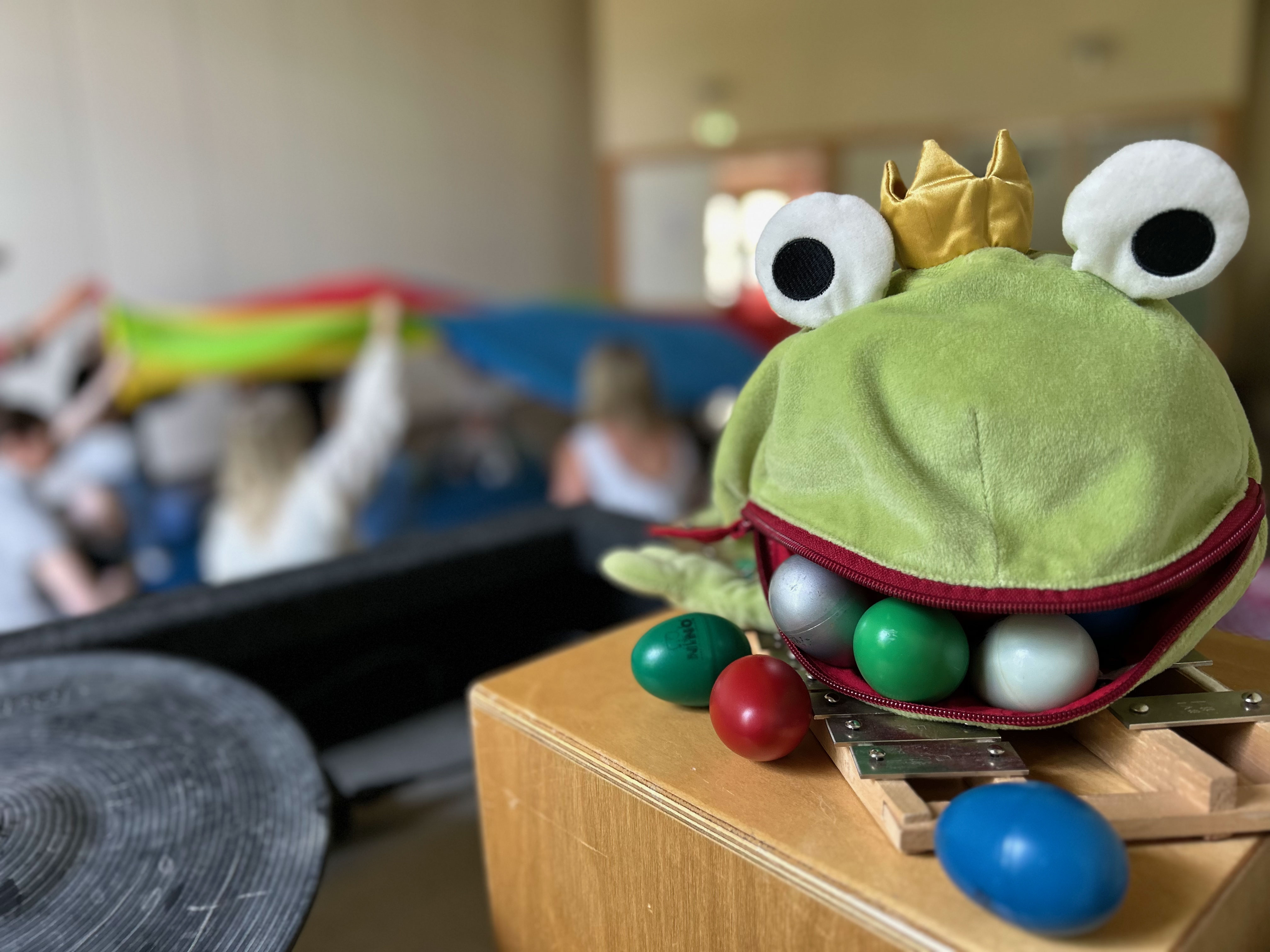 En leksaksgroda full av rytminstrument i förgrunden och föräldrar med bebisar i bakgrunden