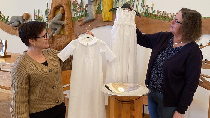 Två dopklänningar i lite större barnstorlekar hålls upp av två medarbetare som står vid en dopfunt.