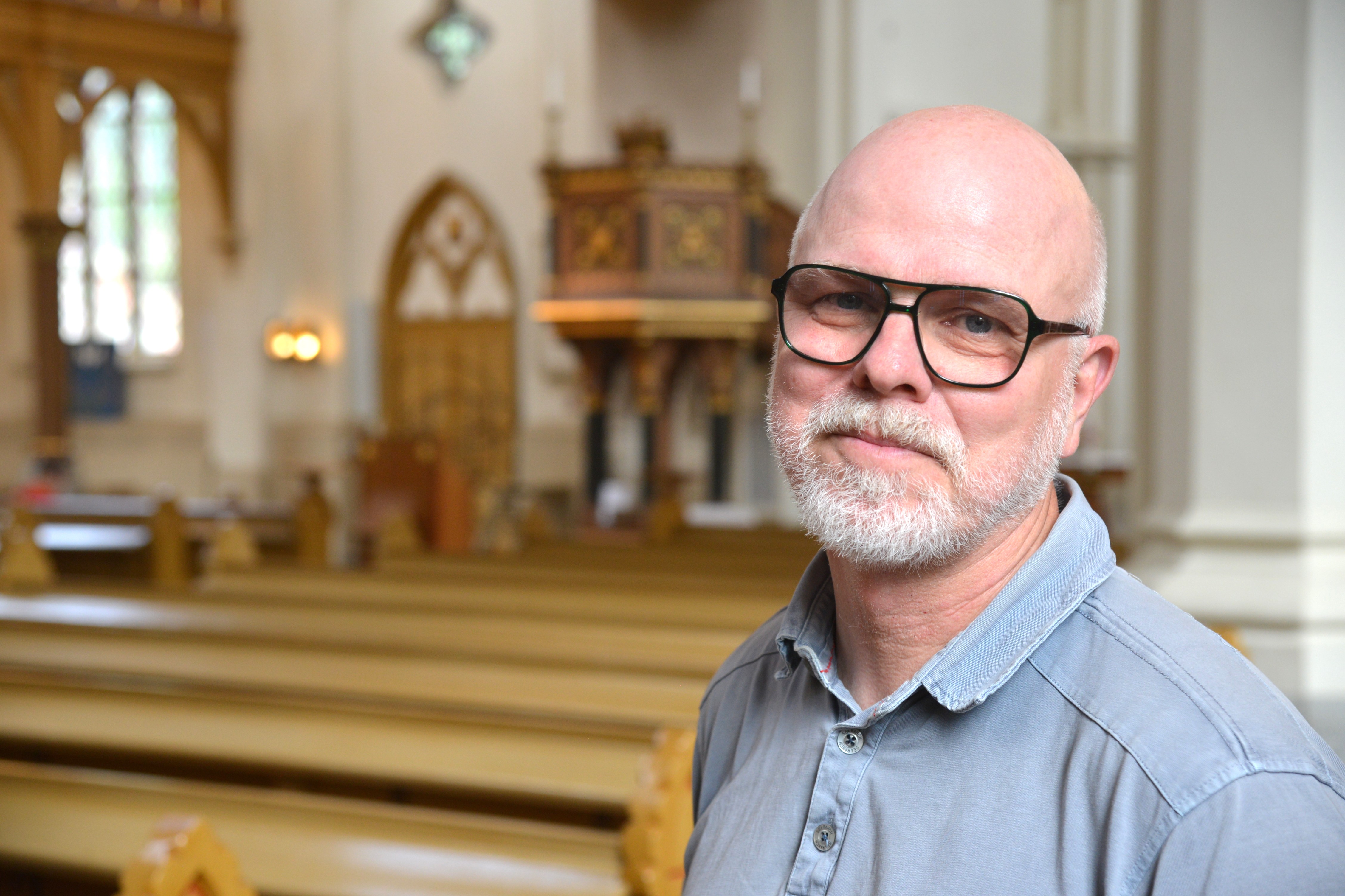 Poerträtt av en man i blå skjorta och glasögon som står i ett kyrkorum
