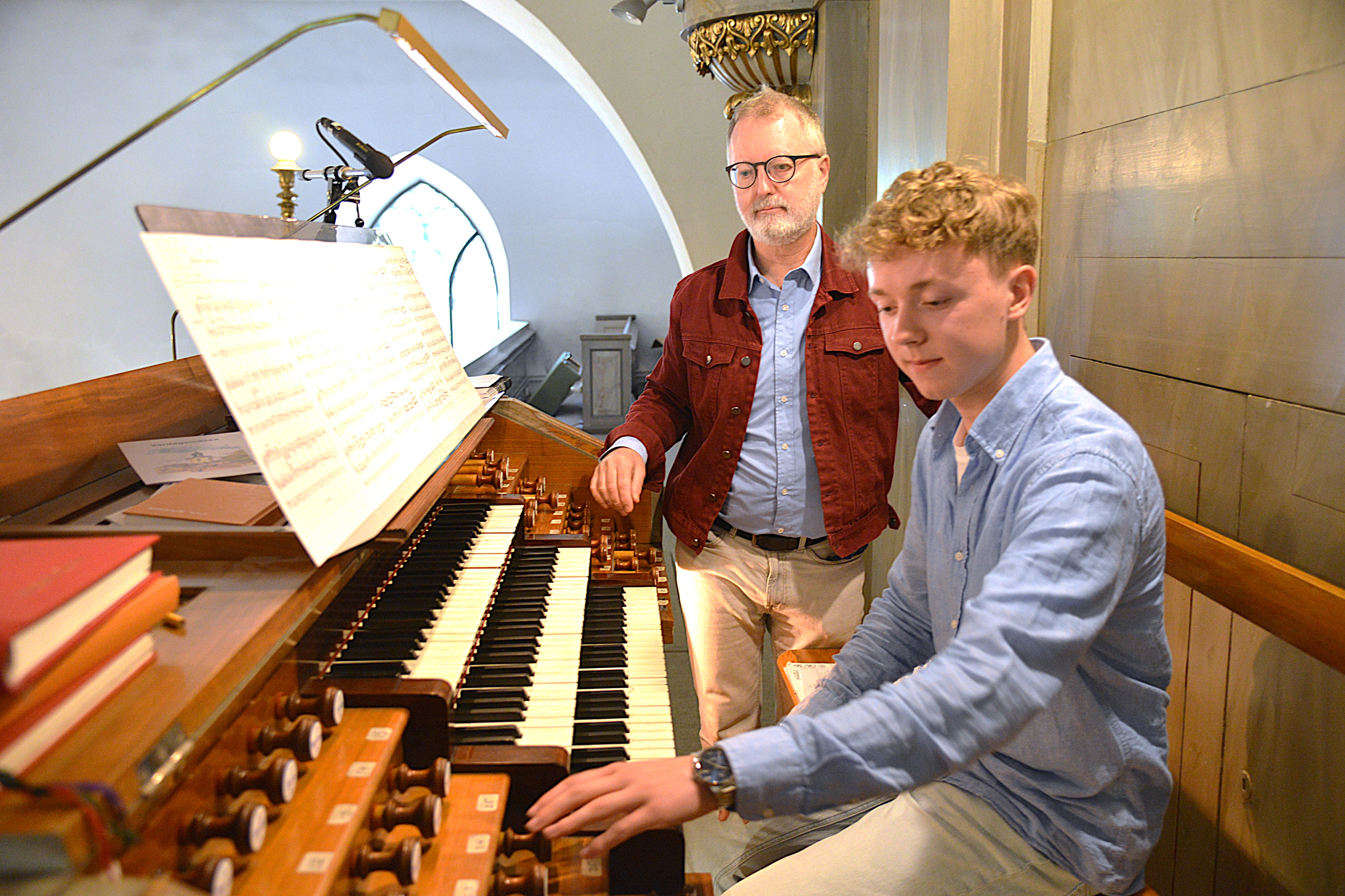 En pojke sitter vid en orgel. En man står intill honom.