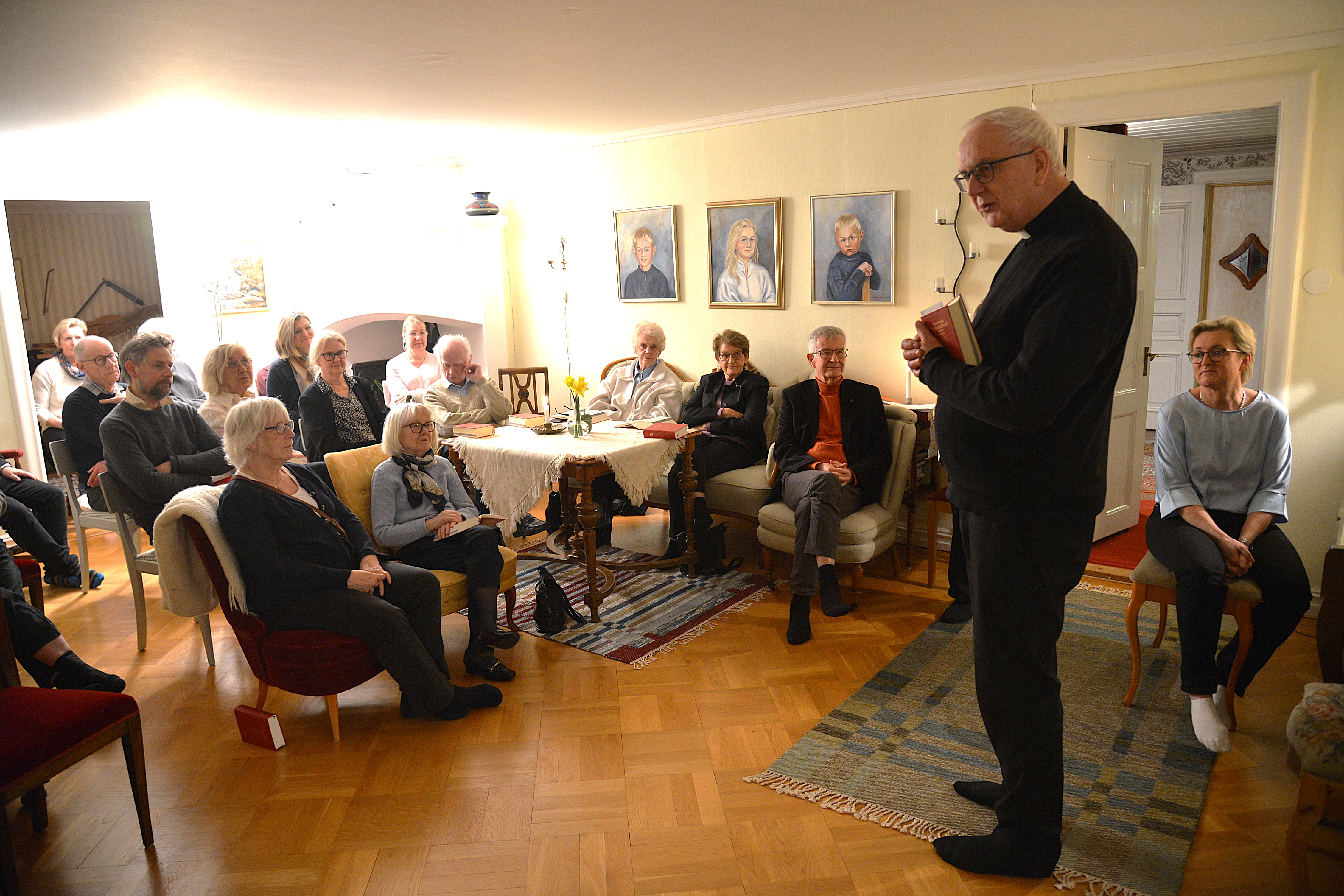 En man i prästkläder står i ett vardagsrum fyllt av människor