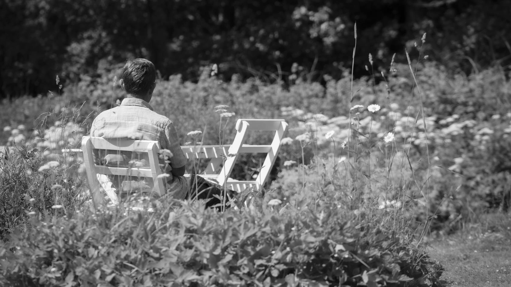 En ensam person sitter i en trädgårdsstol med ryggen vänd mot kameran. Svartvit.
