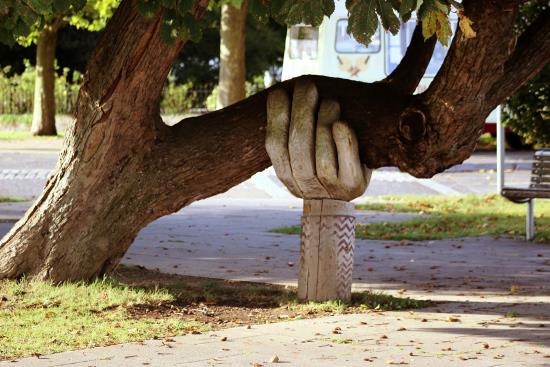 En träskulptur i form av en hand stöttar upp ett stort lutande träd. 