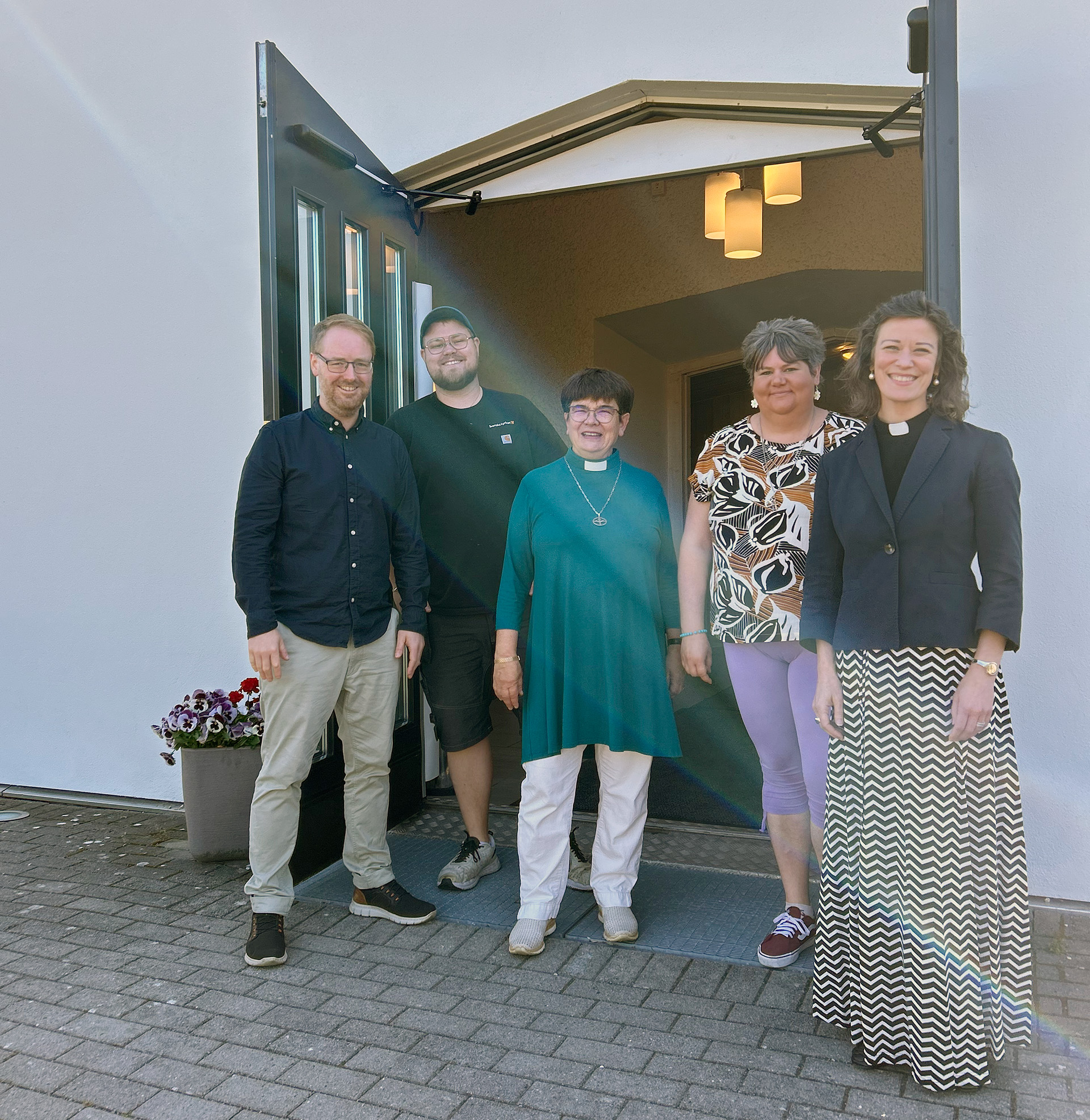 Tomas, Carl, Helena, Julia och församlingsherde Tilda står leende framför den öppna kyrkporten. 