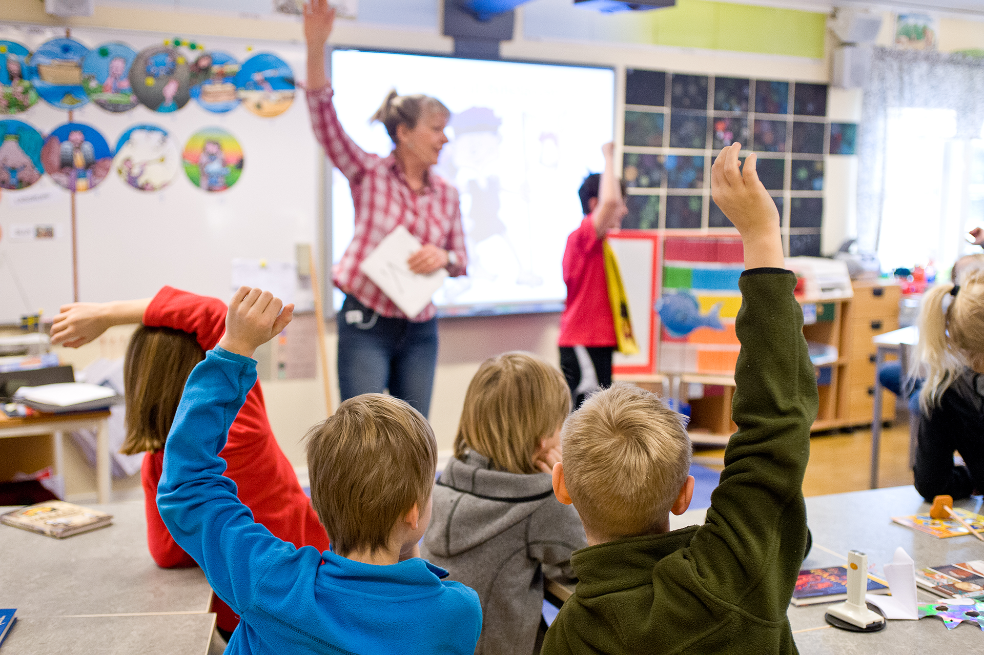 Lärare i bibeläventyret står längst fram i klassrummet. Man ser ryggarna på barn som räcker upp händerna. 