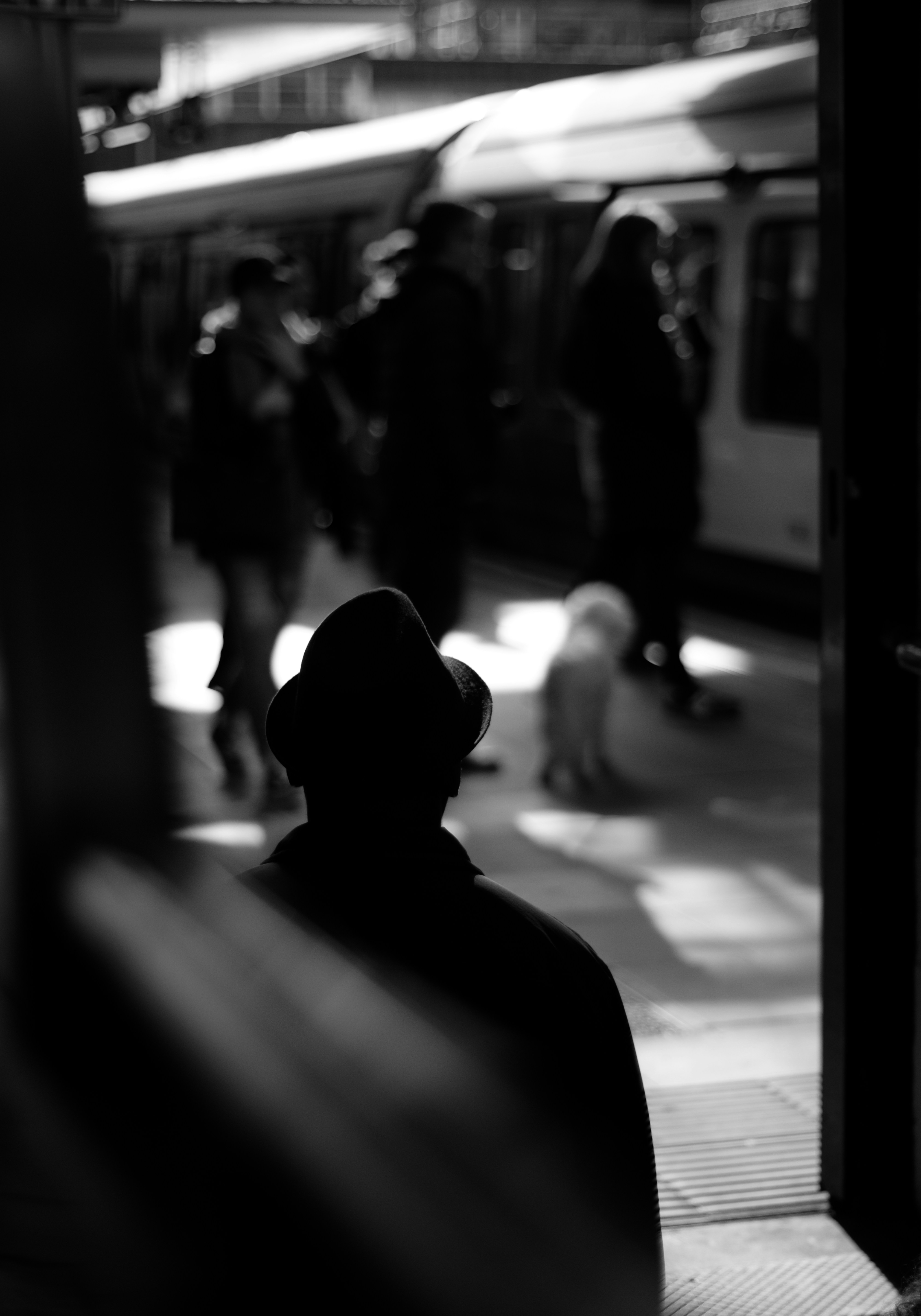 Svartvit bild i silhuett av en man i hatt sedd bakifrån, som blickar ut mot en tågperrong där människor och ett tåg syns i bakgrunden.