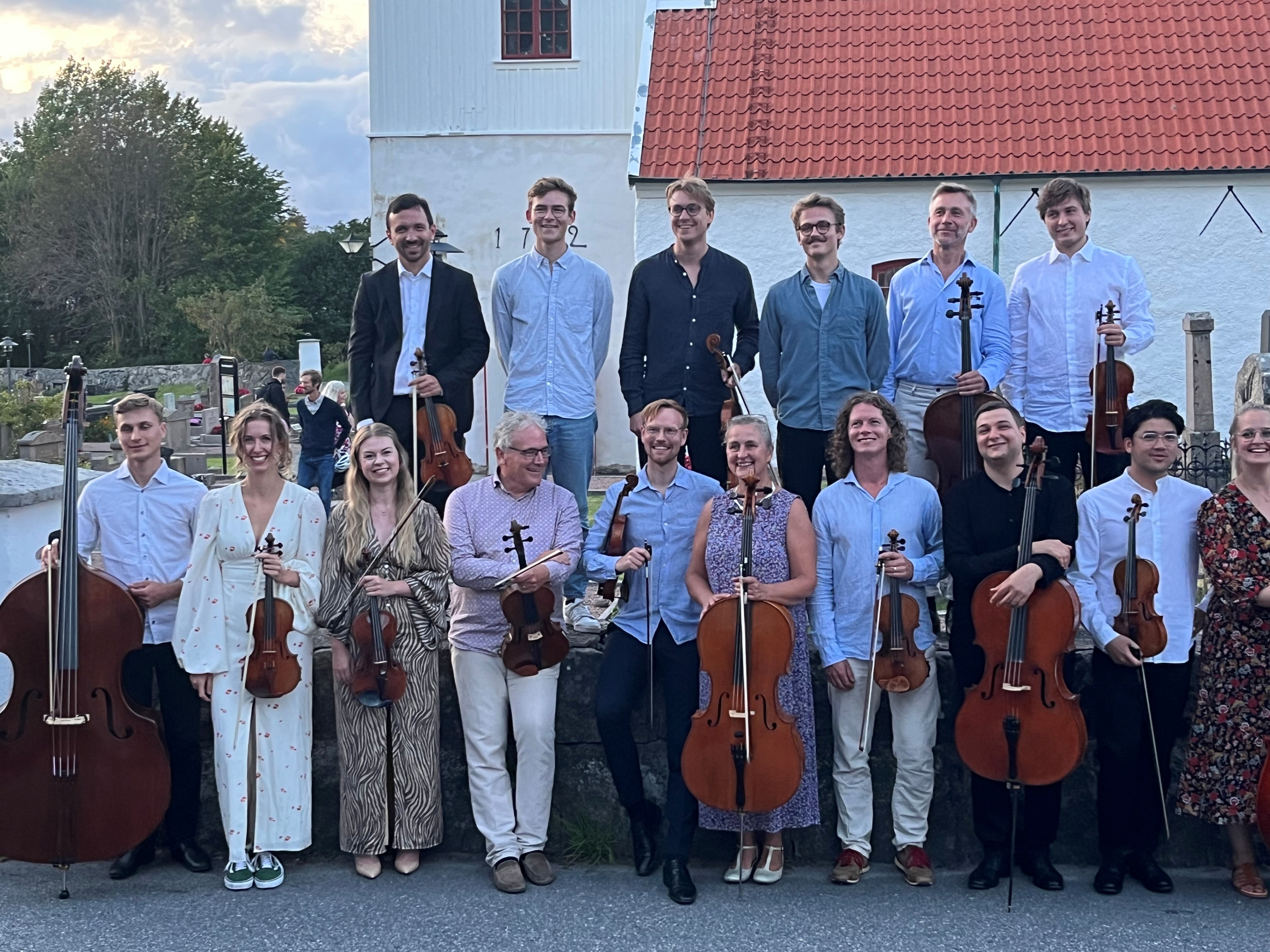 Många musiker i en kammarorkester står uppradade framför en kyrka. De håller i stora och små stråkinstrument och ler mot kameran.