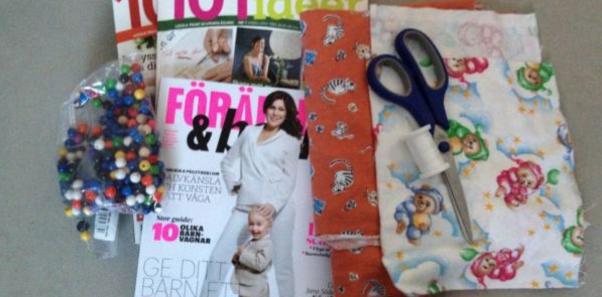 Tråd, sax, plastkulor, tyg och några tidningar ligger på ett bord. 