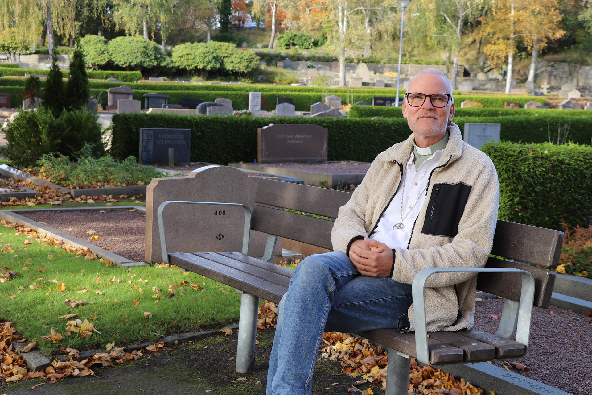 En man i diakonskjorta sitter på en bänk på en kyrkogård. 