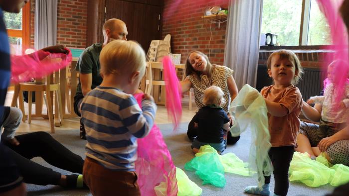 Barn och föräldrar sitter på golvet och leker med färgglada sjalar. 