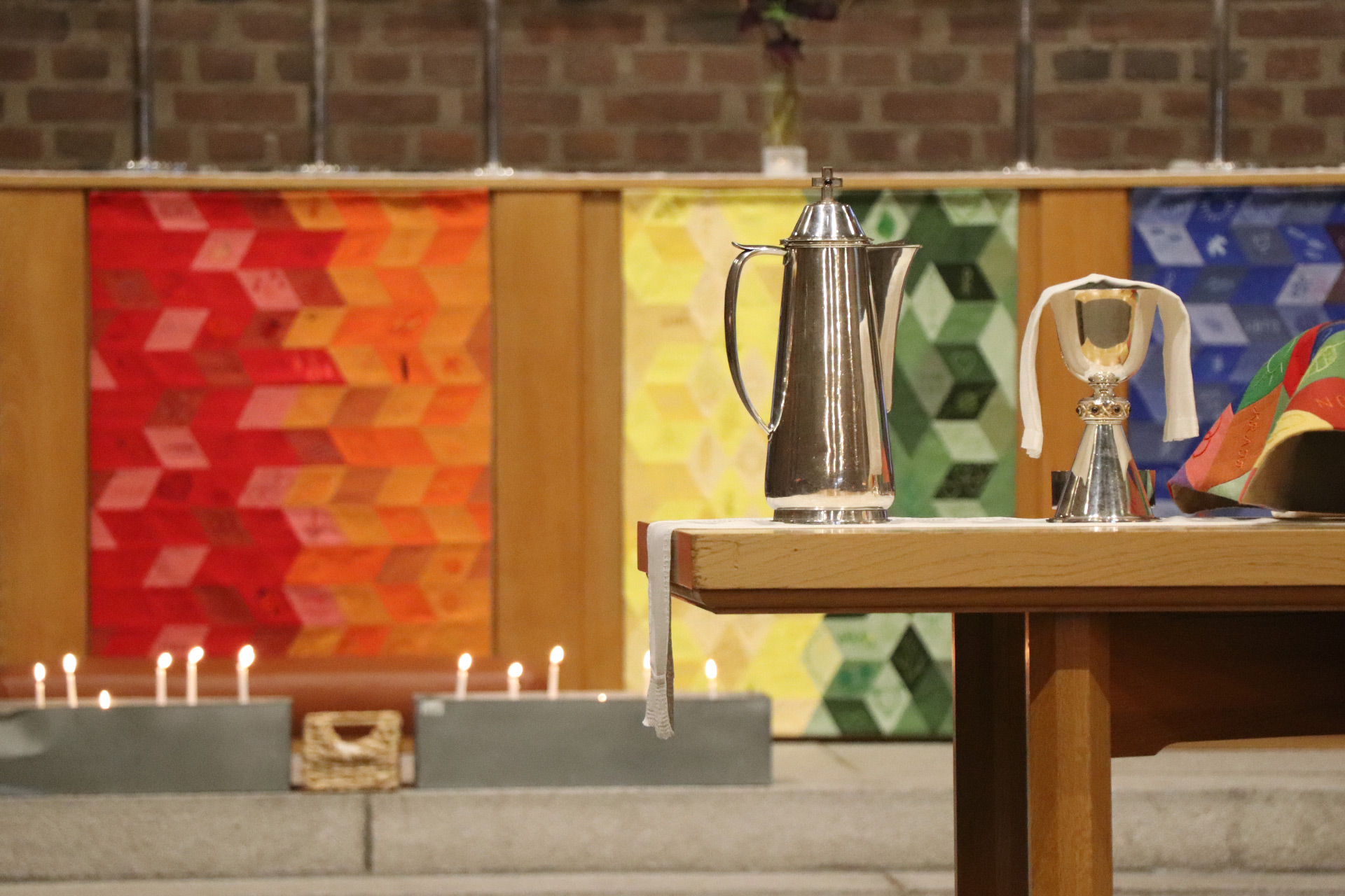 Nattvardskärl på ett bord, i bakgrunden syns altare med altarkläde i regnbågens färger. 