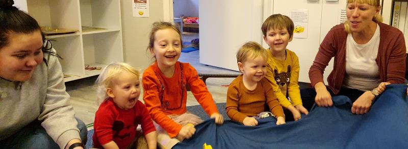 Barn och vuxna sitter på golvet, håller ett blått tygstycke mellan sig och skrattar.