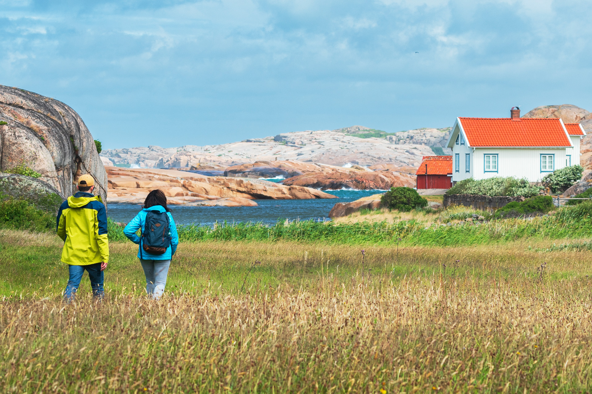 Två personer promenerar i havsnära landskap med klippor, hav och några hus.