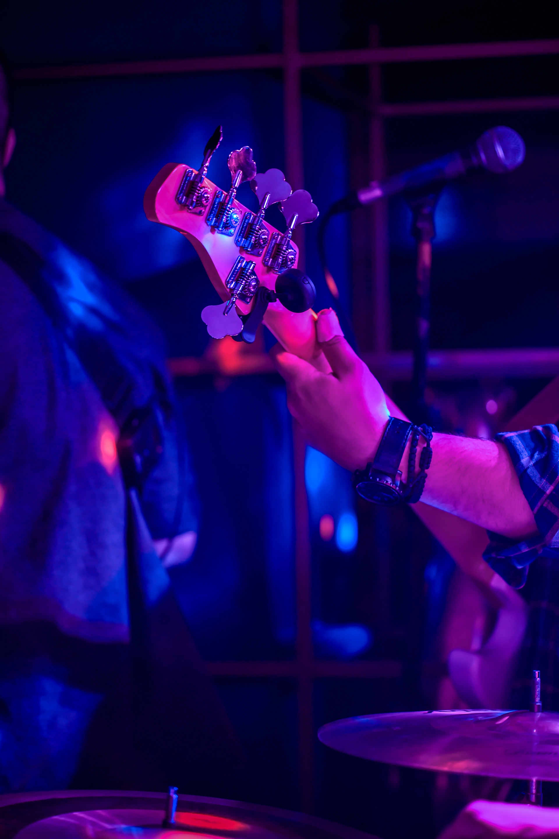 Närbild av en basgitarrs huvud som spelas på en scen upplyst av lila och blått ljus; i bakgrunden syns suddigt en mikrofon och ett trumset.