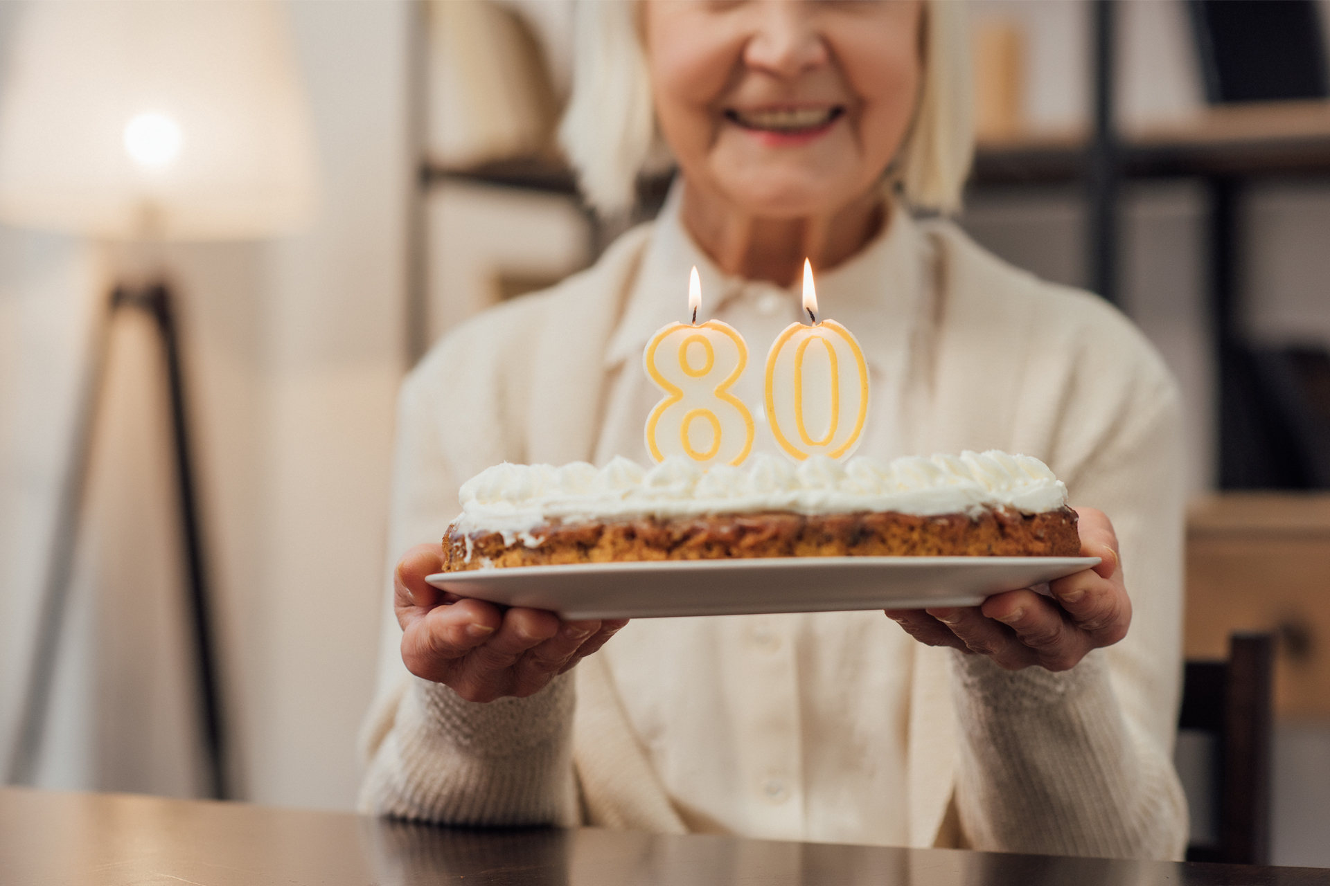 Äldre kvinna håller upp födelsedagstårta. På tårtan finns ljus som formar siffran 80.