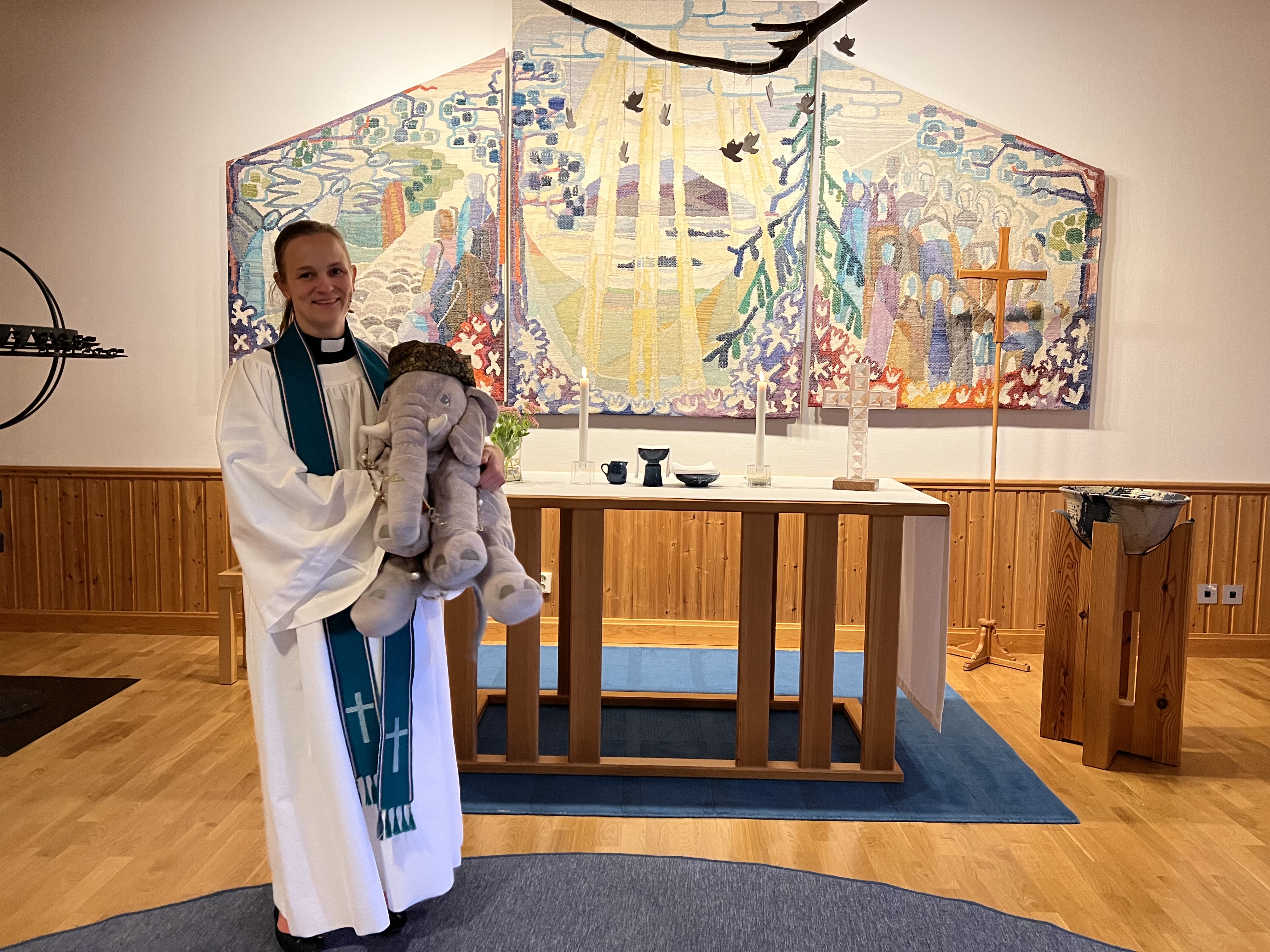Prästen Karin och elefanten Zambezi (en grå plyschelefant) vid altaret på Mikaelsgården.en