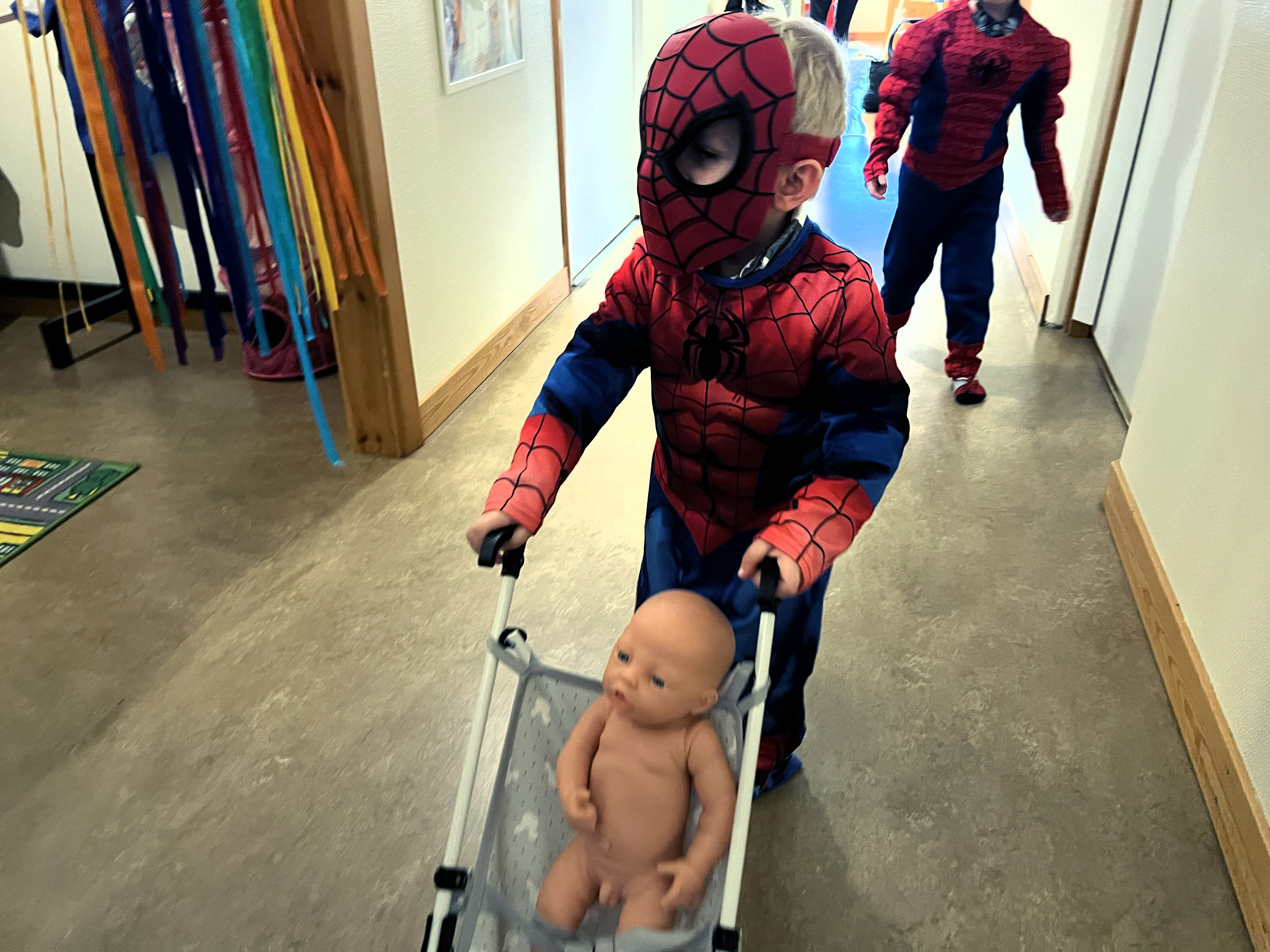 Ett barn i spindelmannendräkt drar en docka i en dockvagn.