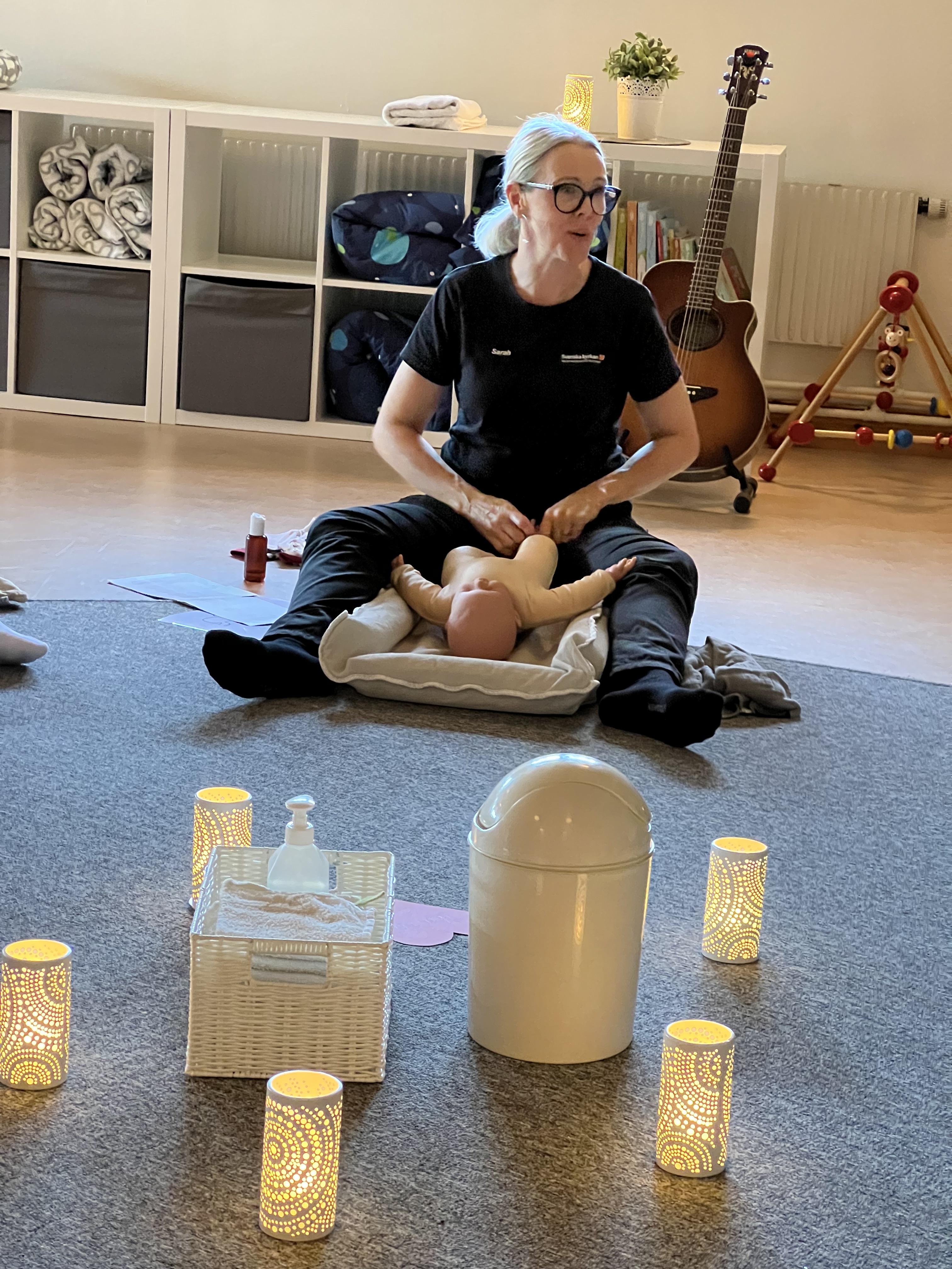 En kvinna som sitter på golvet och visar på en docka hur man masserar sitt barn.