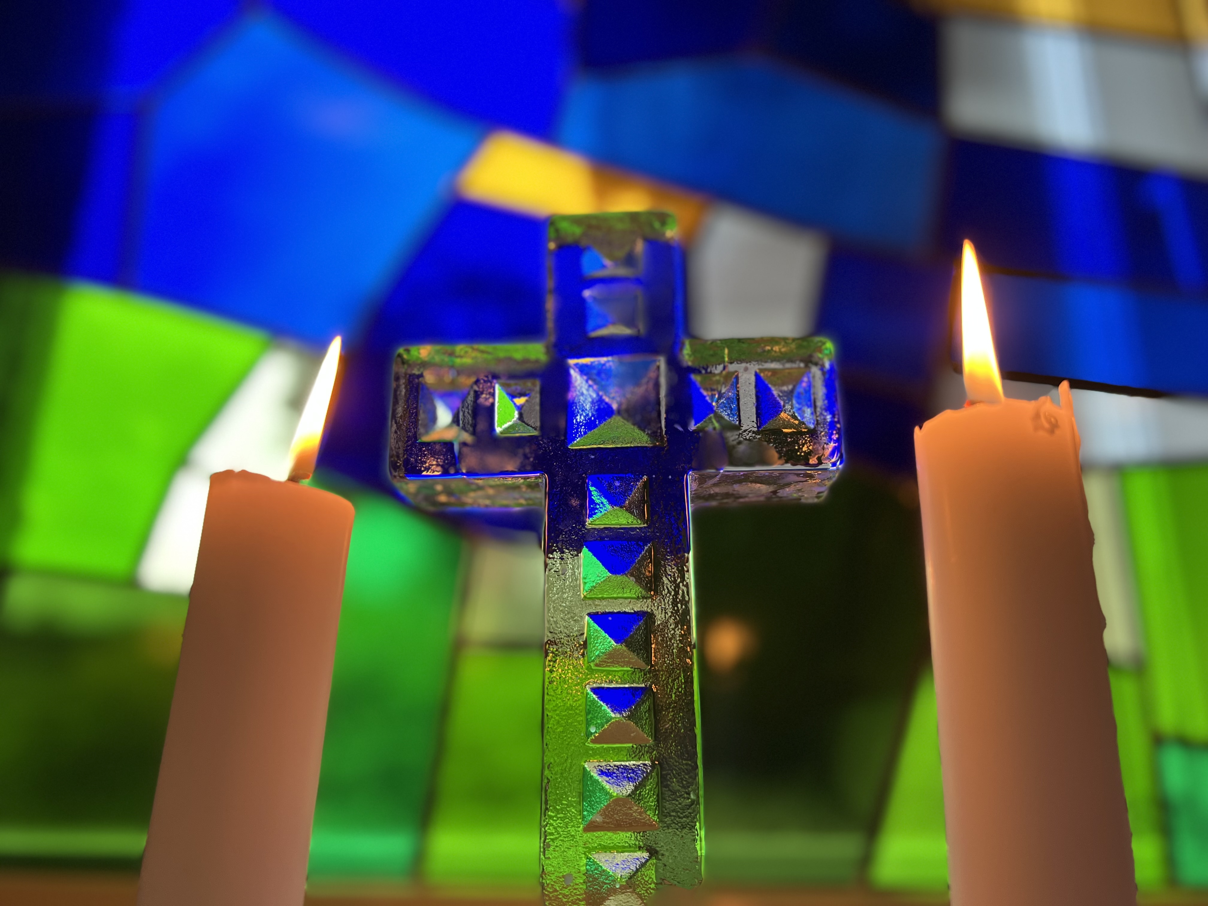 Två tända ljus och ett glaskors vid ett blått kyrkfönster.