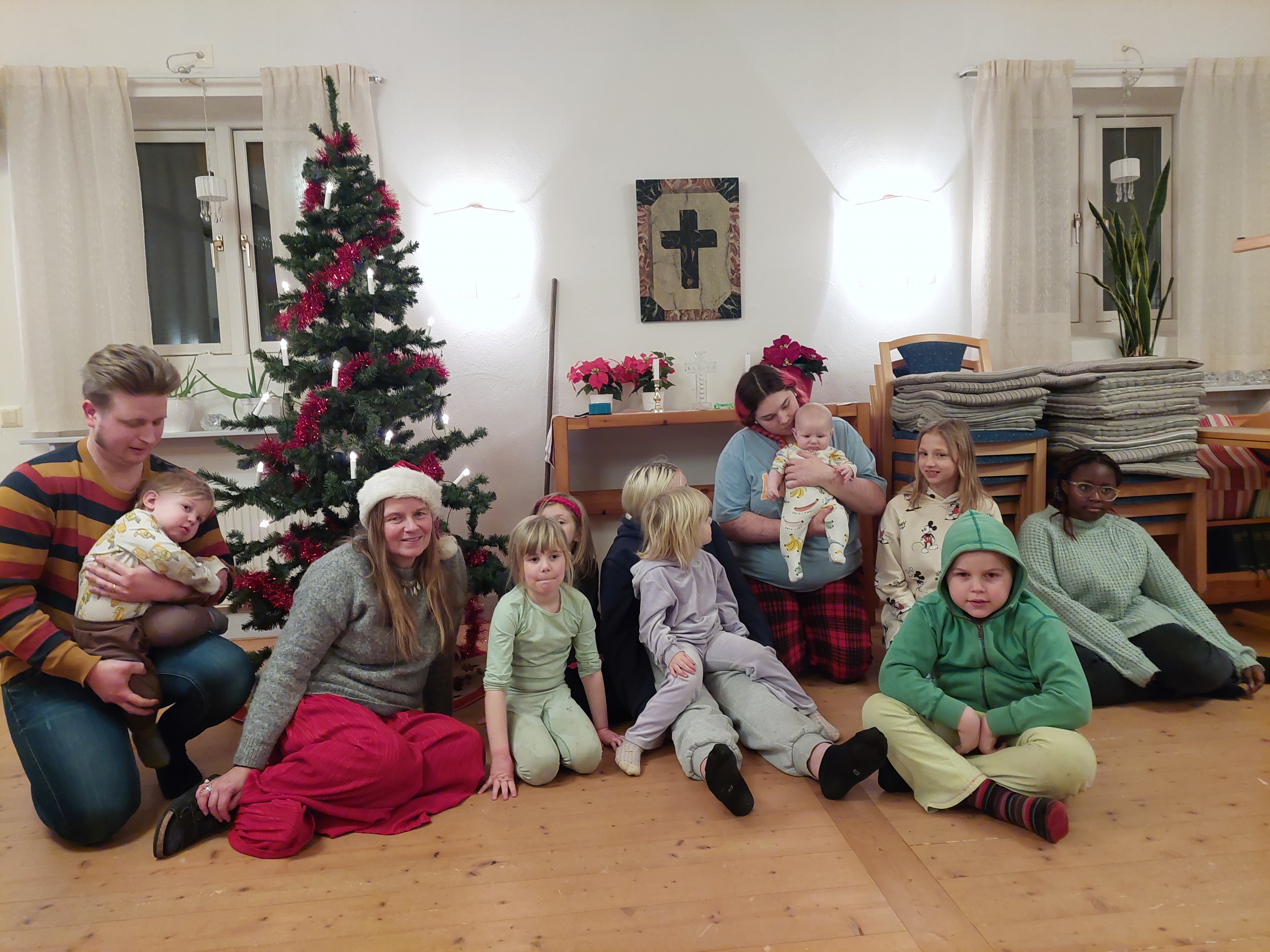 En grupp med vuxna och barn sitter vid fotan av en pyntad julgran i Dals sockenstuga