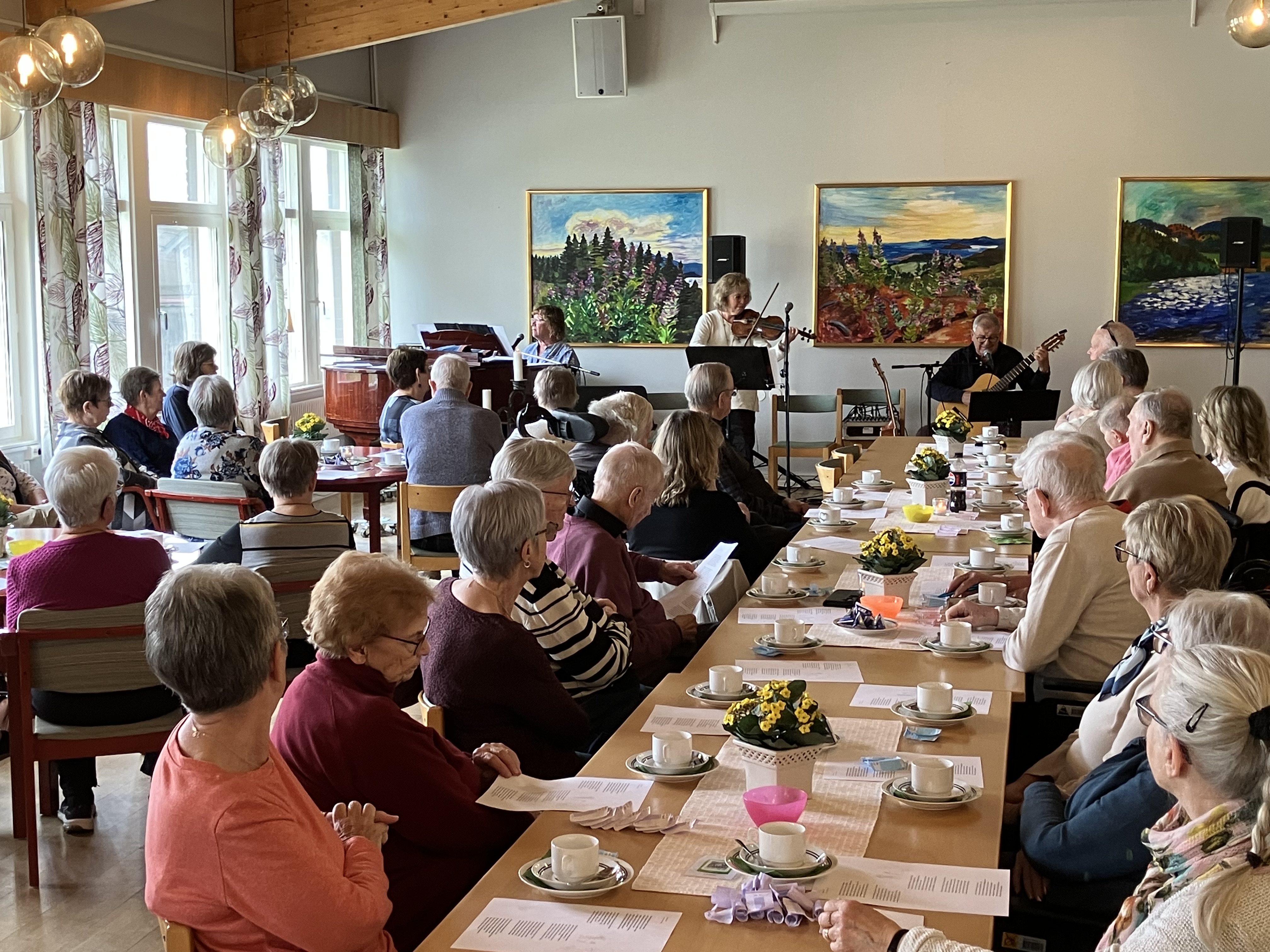 Äldre människor samlade runt fikabord lyssnandes till livemusik
